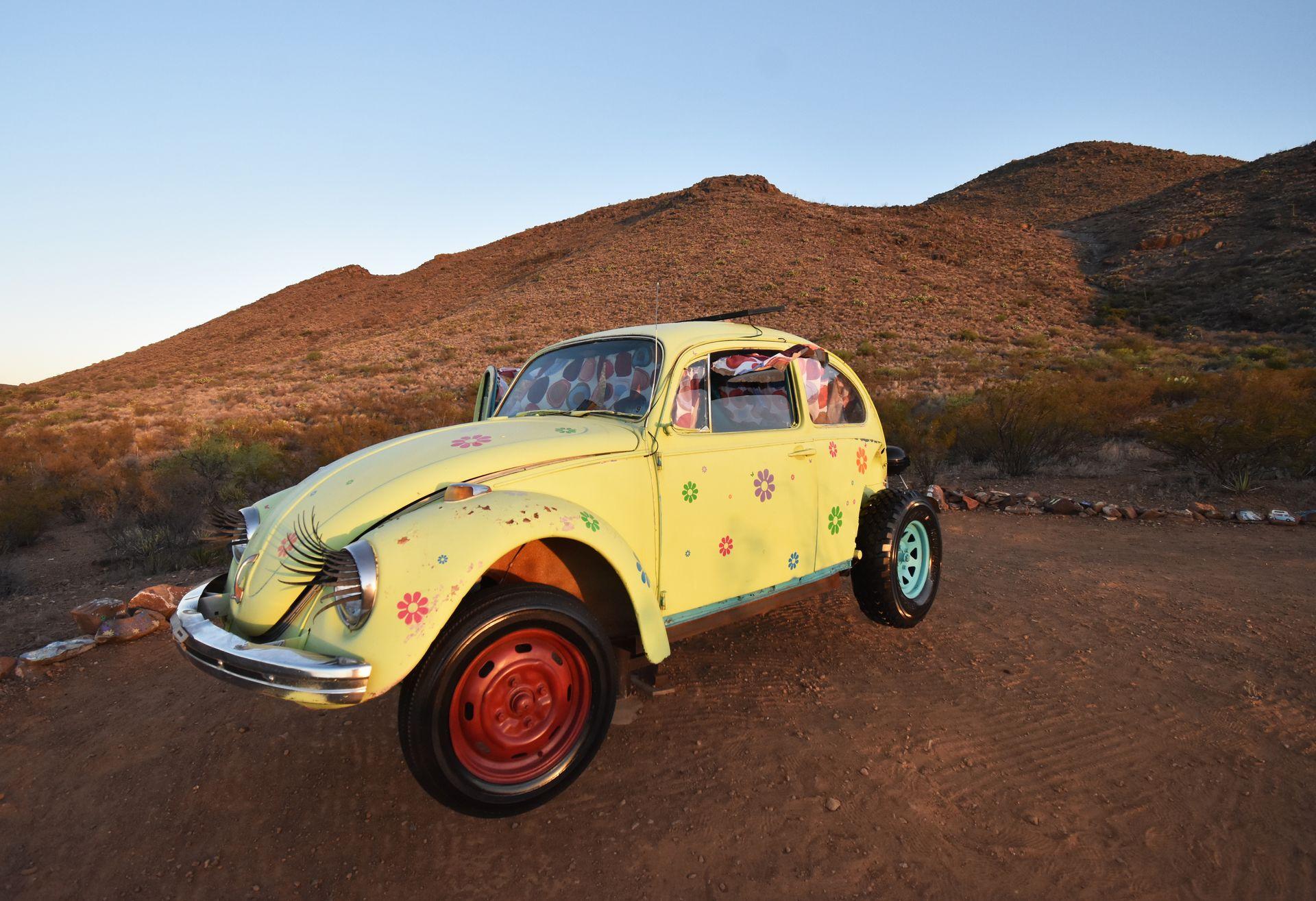 A yellow Volkswagen beetle elevated by a rod with mountains in the background. The bug has eyelashes and some colorful flowers on it.