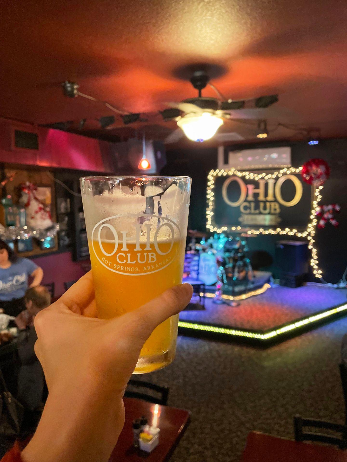 Holding up a beer inside of the Ohio Club. There is a stage for live music in the background.