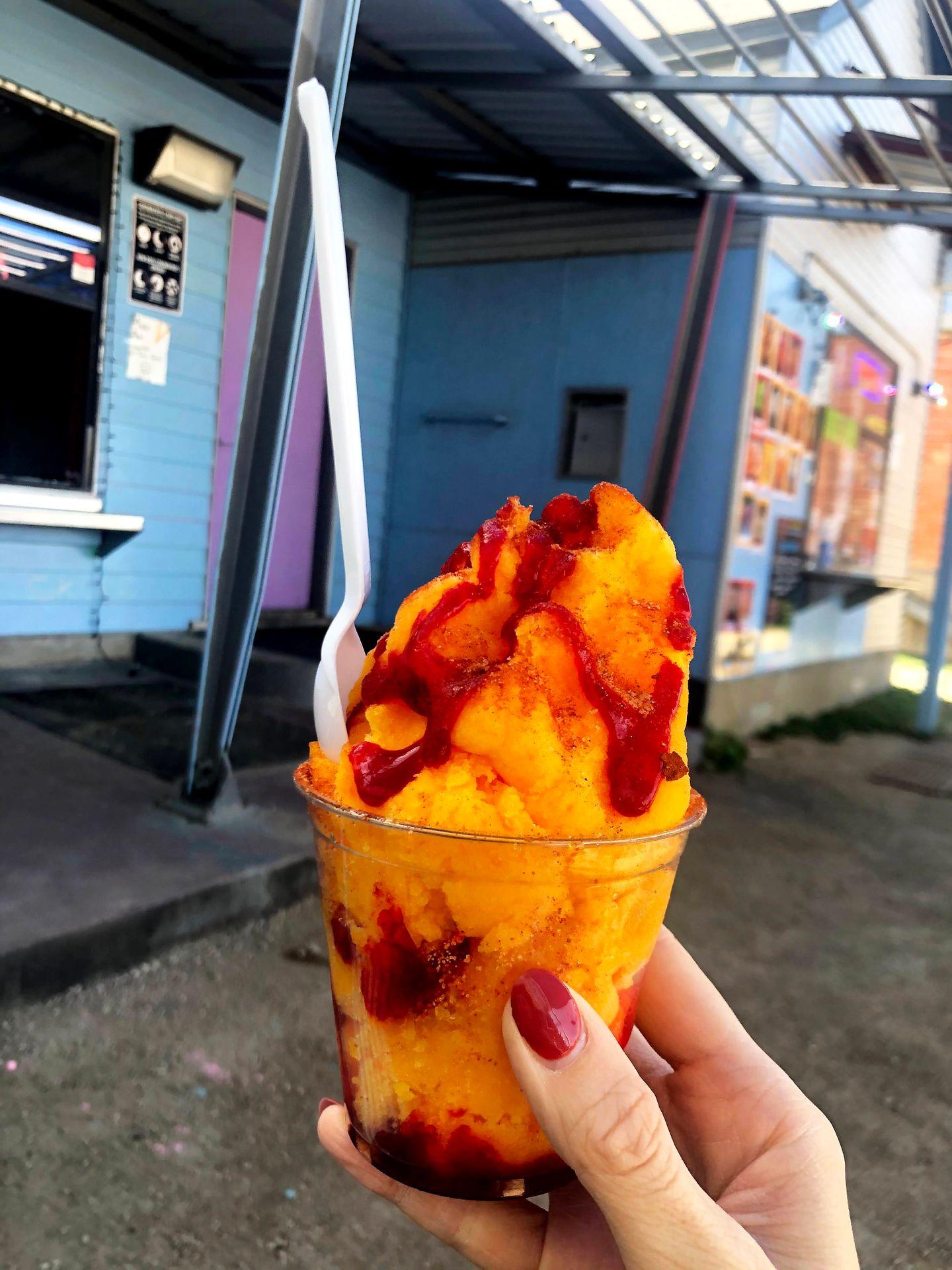 Holding up a chamoyada. There is mango shaved ice topped with red chamoy sauce.