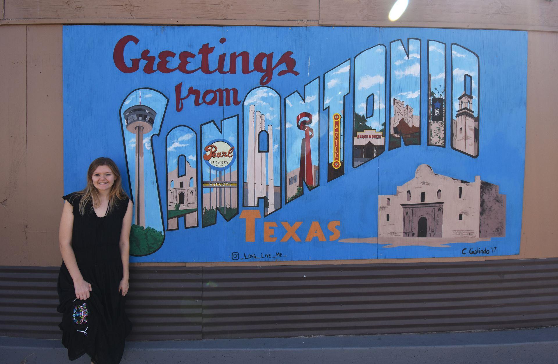 Lydia leans up against a mural that reads 'Greetings from San Antonio Texas'