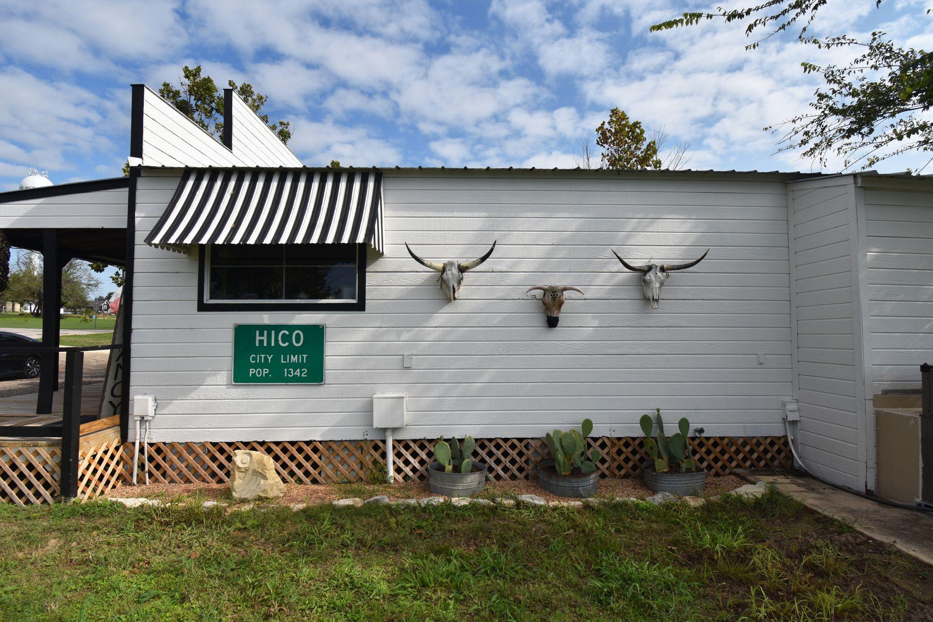 A white building with 3 Texas longhorn skulls and a sign reading "Hico City Limit: Pop. 1342"
