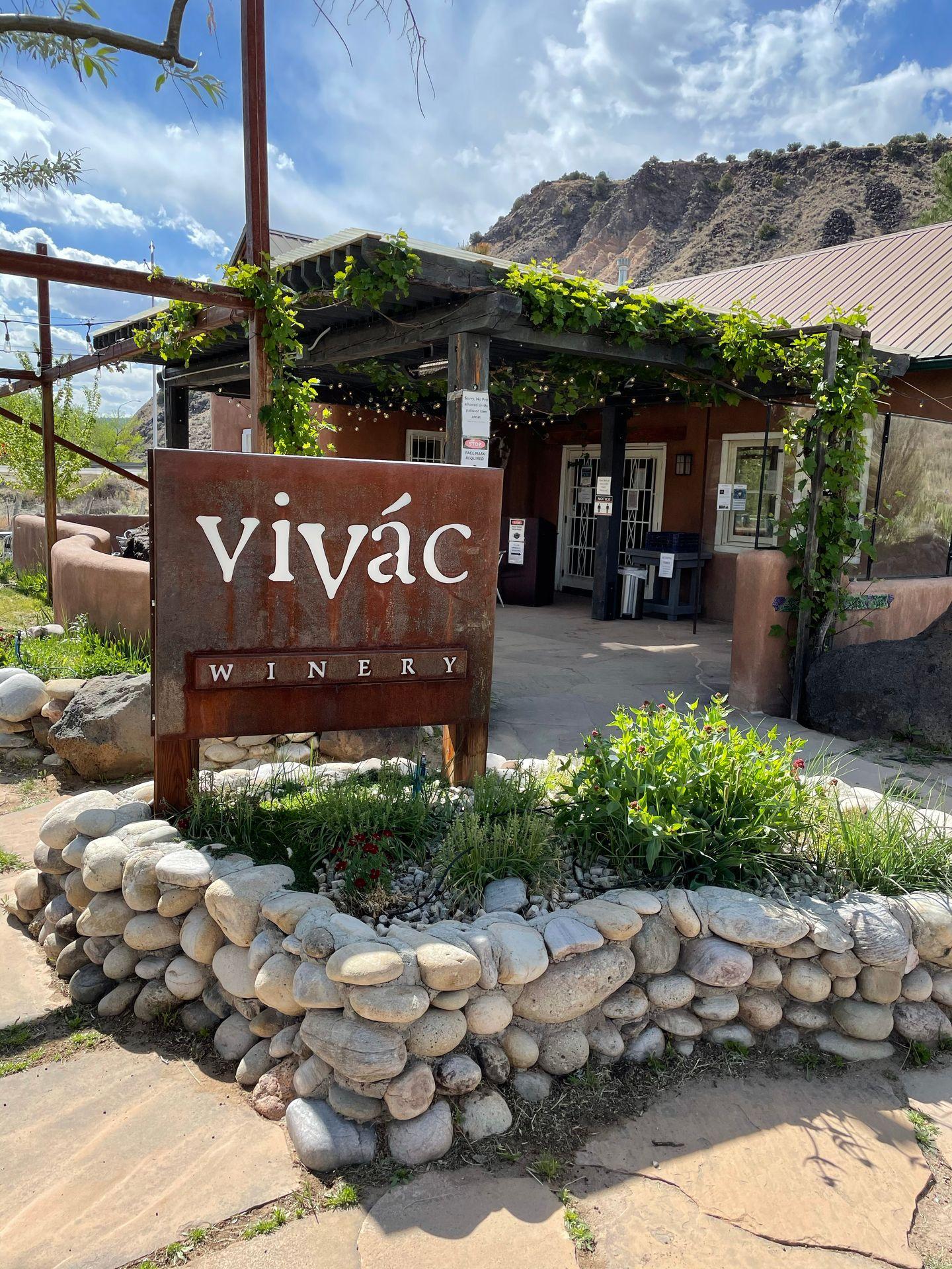 The exterior of Vivac Winery. There is a sign, greenery behind a stone wall and an overhang with hanging green vines.