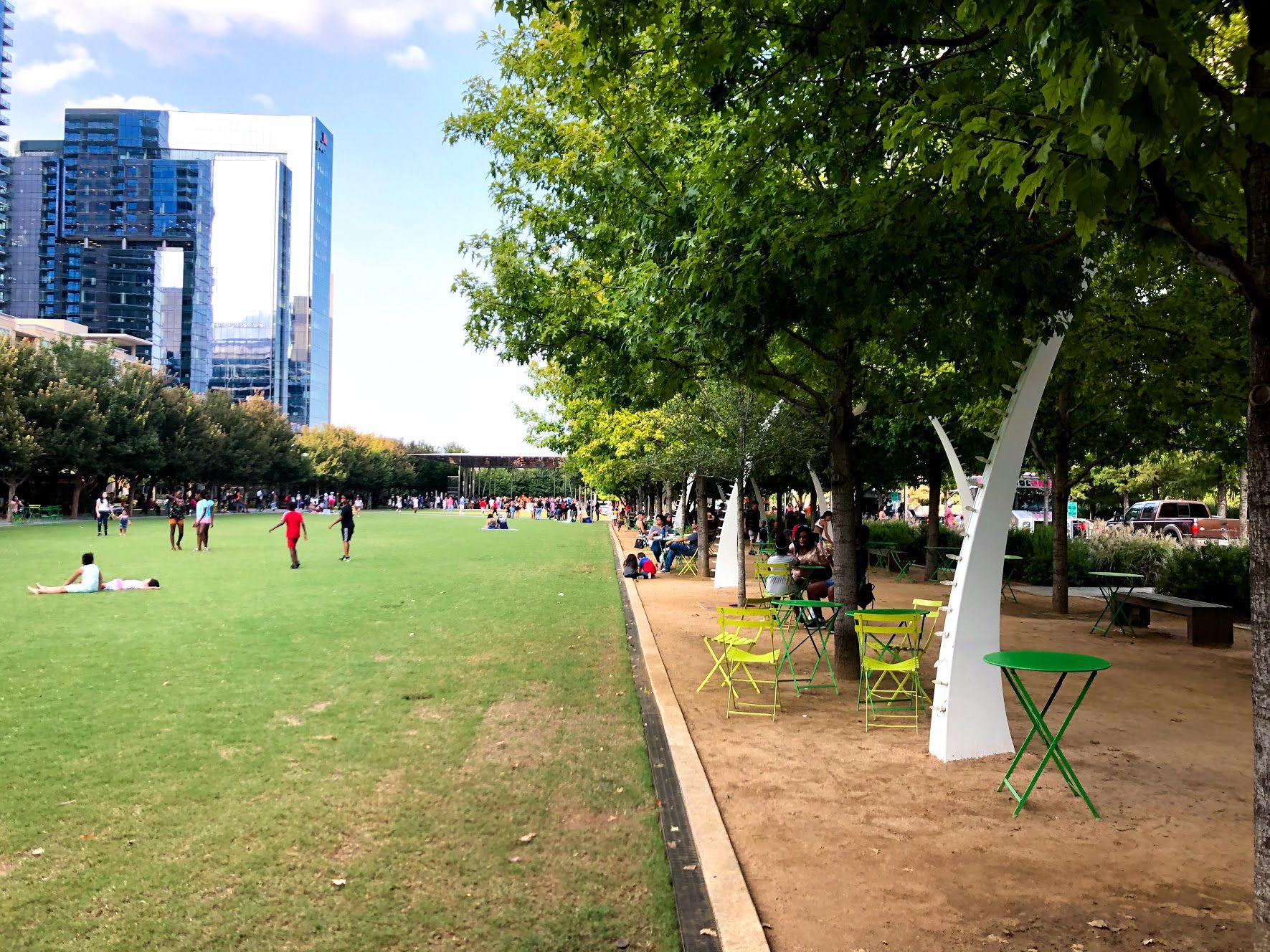 A lawn and a row of arches at Klyde Warren Park. There are small tables with chairs, benches and people walking across the lawn