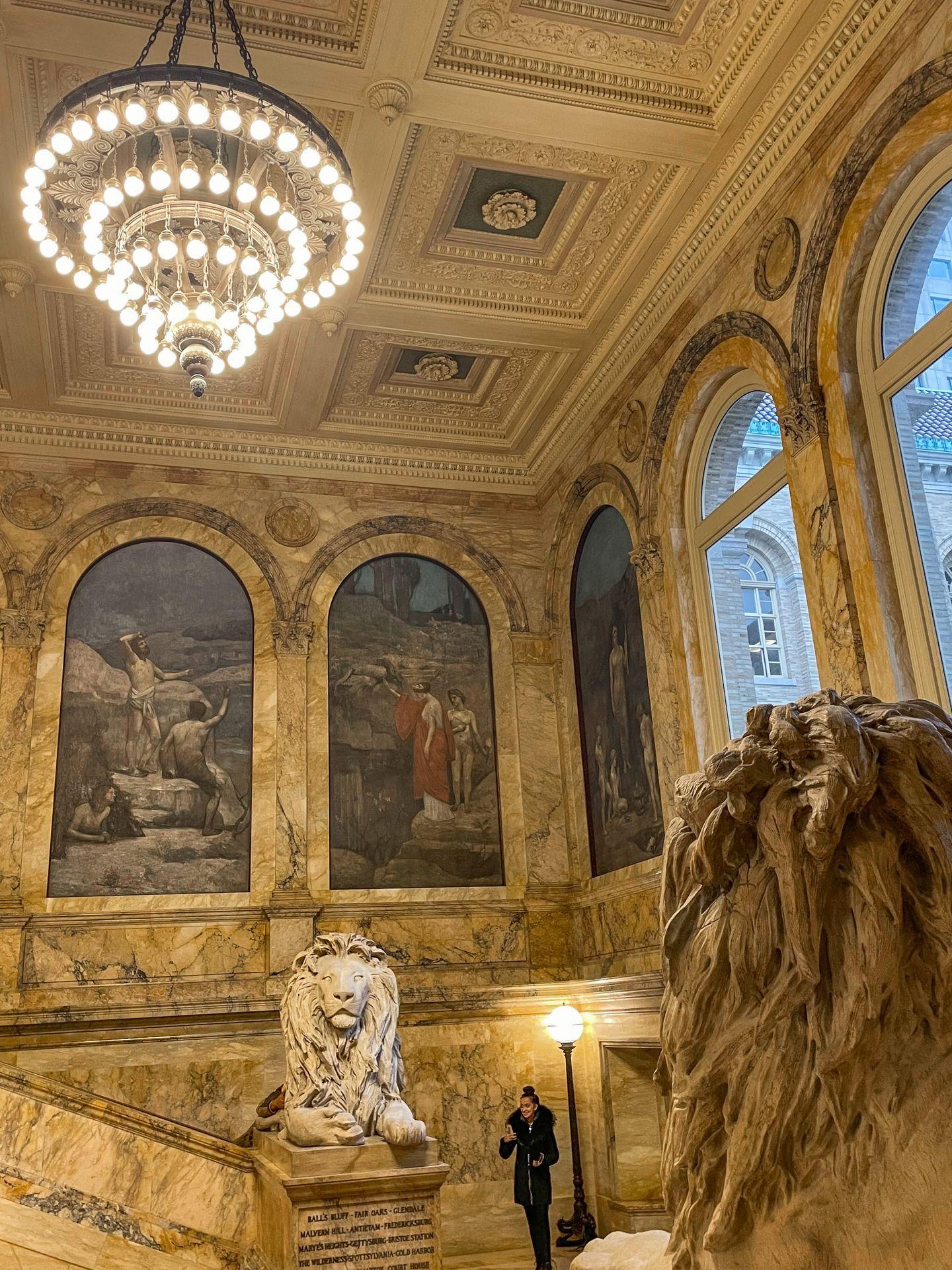 A staircase inside the Boston Public Library that includes artwork, a chandelier and lion statues.