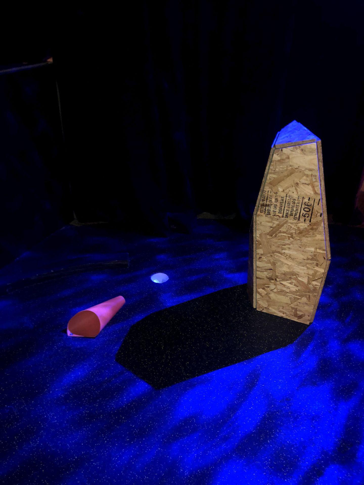 A dark room with blue lights and an abtract-shaped wood monolith at Another Round.