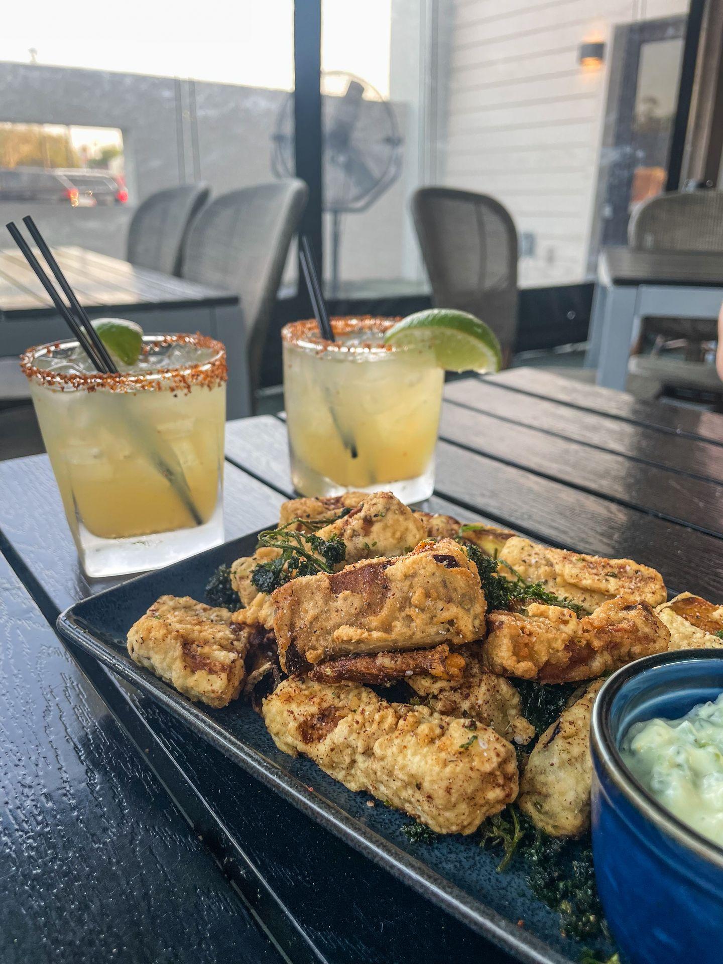 Two margarita's next to an appetizer of fried eggplant and artichokes.