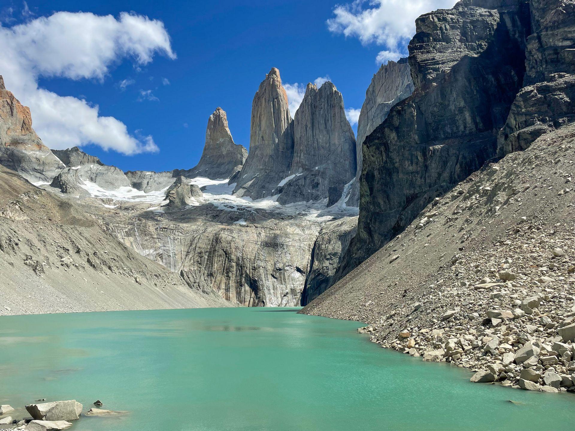 A view of Mirador Las Torres on the W Trek in Patagonia. The view features 3 giant pillars of rock towering in the area with a lake in the foreground.