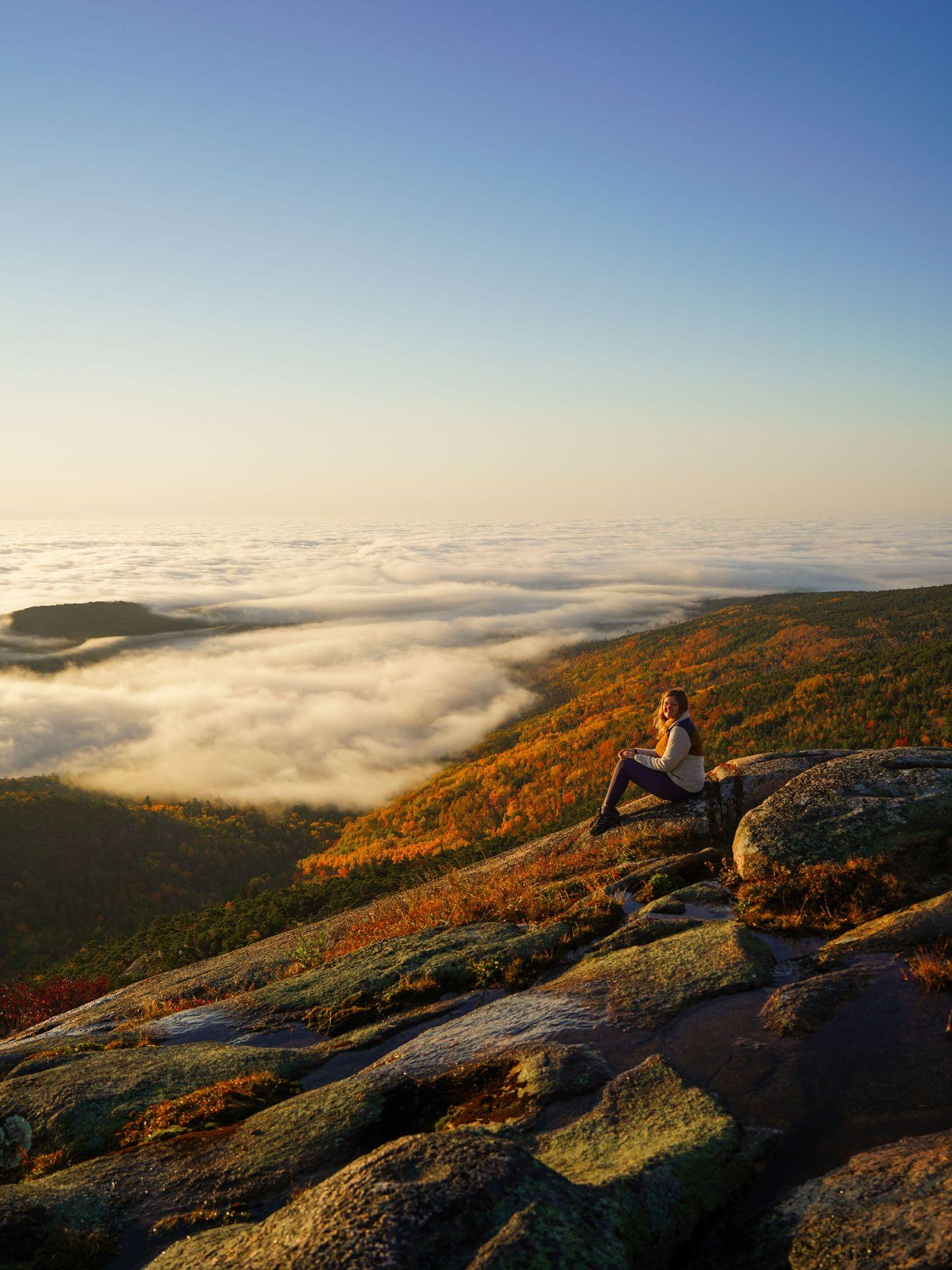 Lydia sitting and looking out at a sunrise with a cloud inversion from Cadillac Mountain