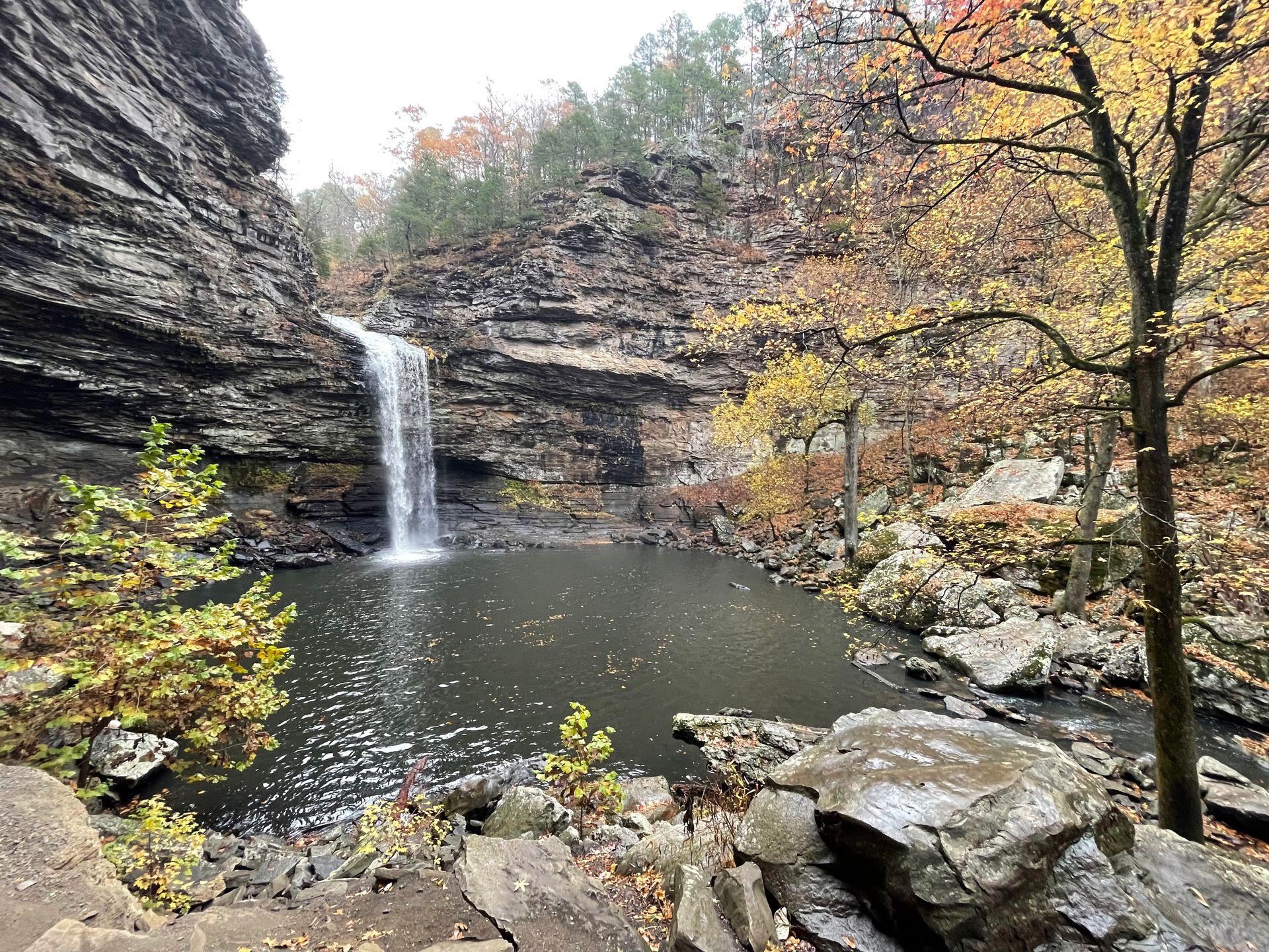 A view of the base of Cedar Falls from across a pool of water. There are some trees with yellow foliage on either side.