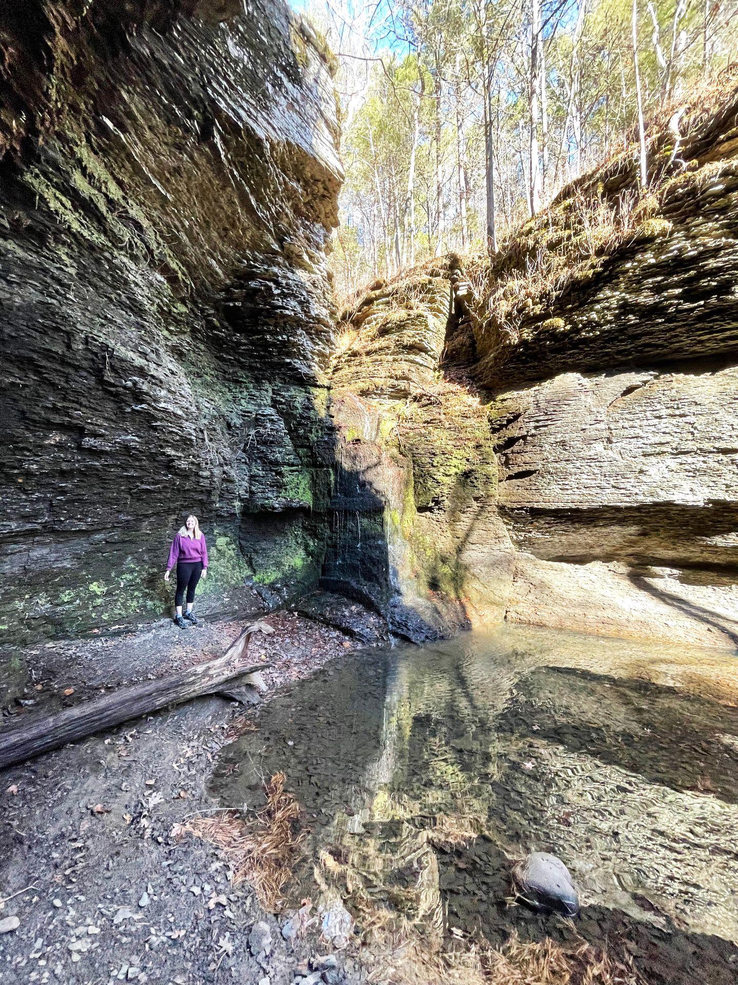 Lydia stands next to a rock wall with Fuzzy Butt Falls to the right. The waterfall is just a trickle.