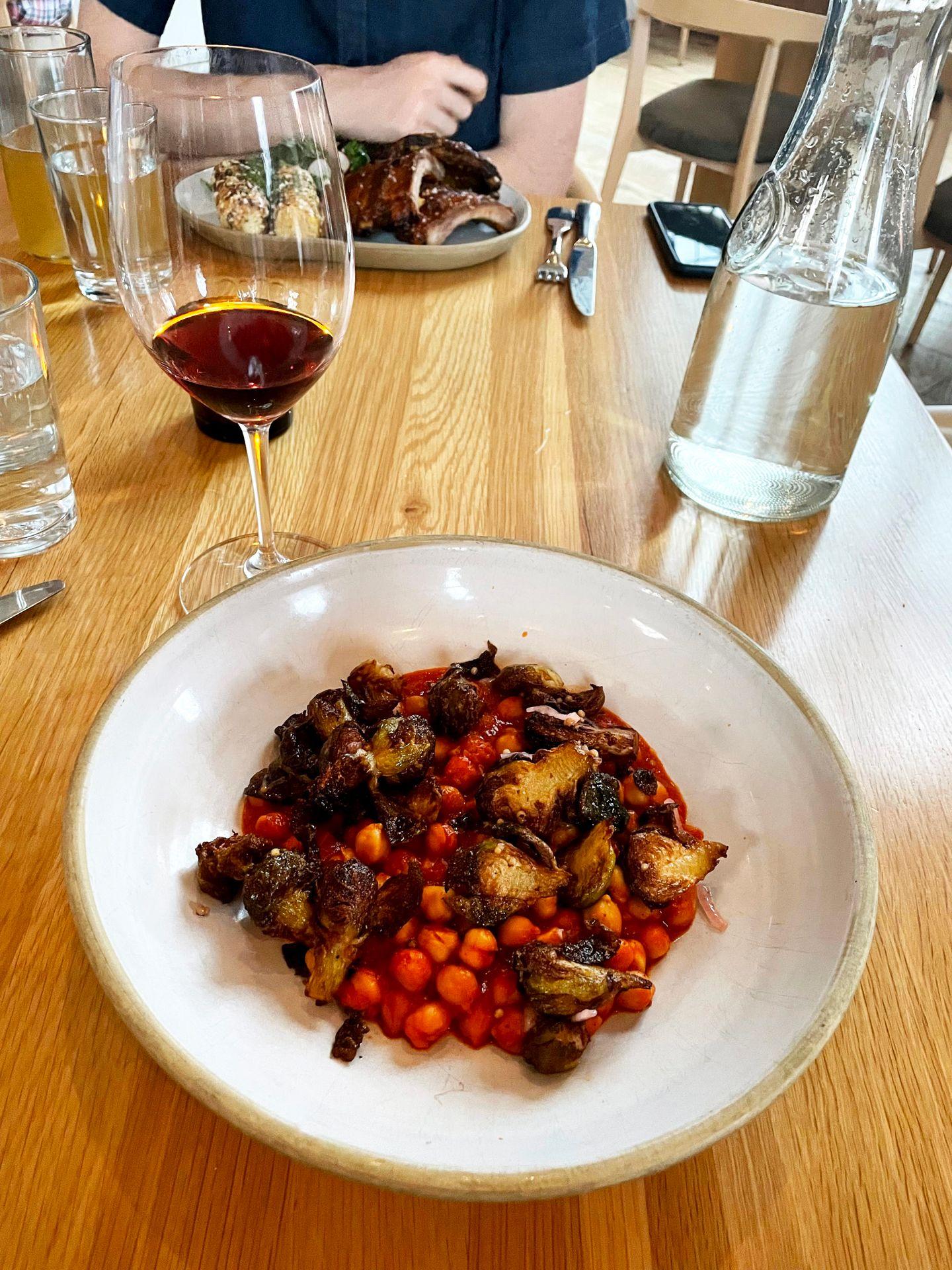 The vegetarian smoked branzino dish. There are chickpeas and brussels sprouts in a red sauce on a white plate. There is also a glass of red wine on the table.