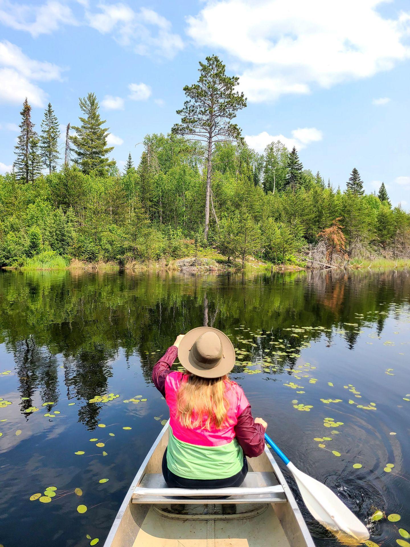 Lydia sitting at the front of a canoe and paddling towards the shore, with some lily pads near to the boat