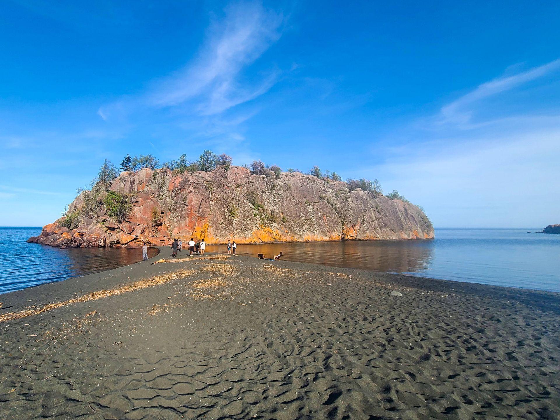 A black sand beach that gets narrow as it extends out to a giant rock formation that is reddish in color.