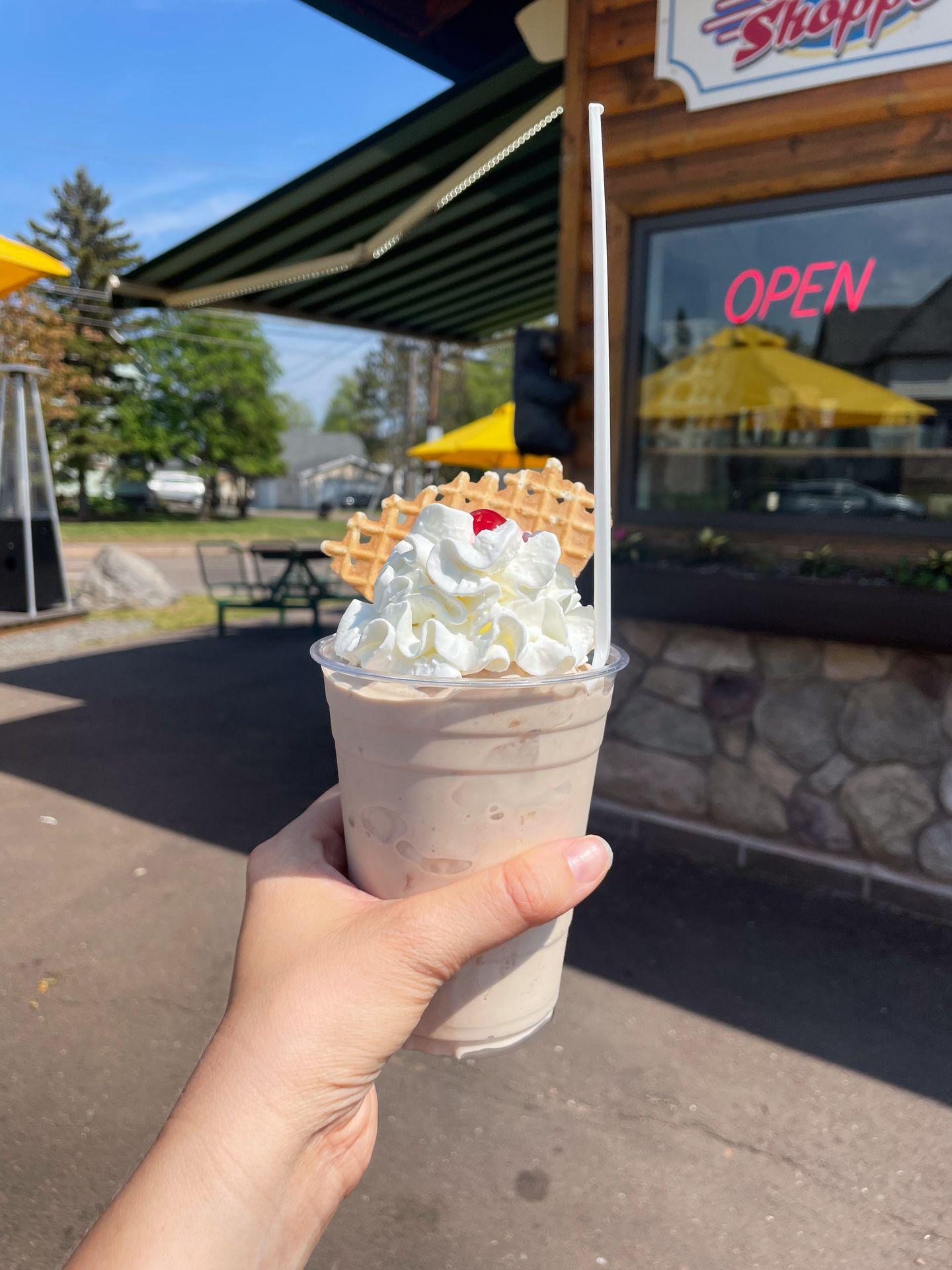 Holding up a chocolate malt topped with whipped cream and a cherry in front of the 5th Street Malt Shoppe.