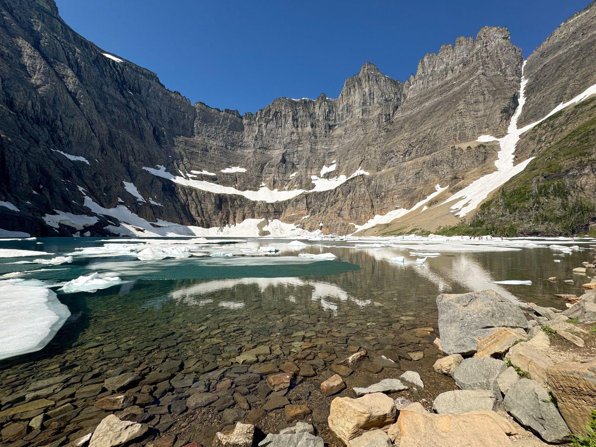 A blue lake with icebergs floating around. There are mountains on the opposite side of the shore.