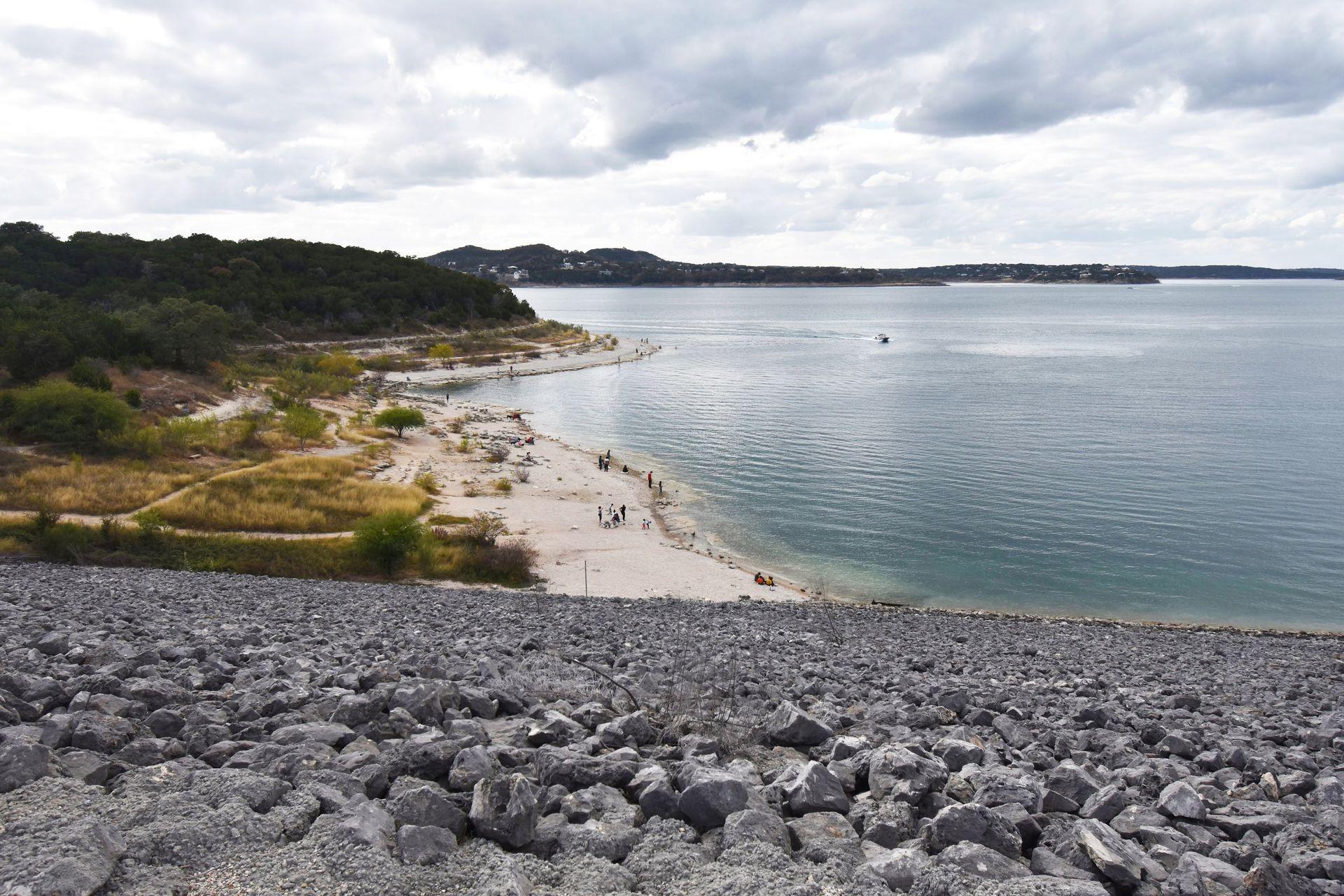 Looking down from a dam at a blue lake and a shoreline with a beach and trees.