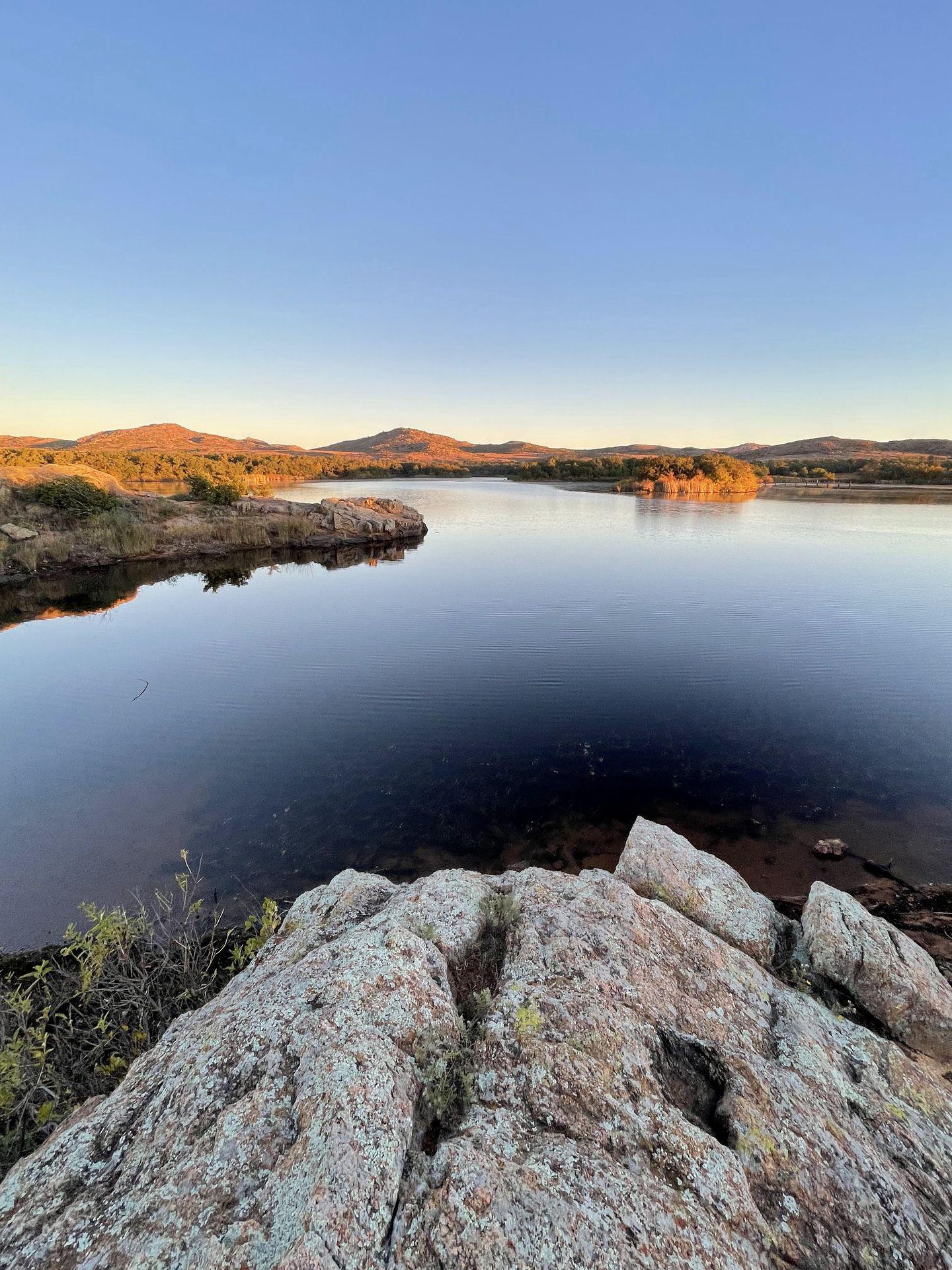 A lake in the Wichita Moutains. Across the water, the hills and trees are glowing as the sun rises.