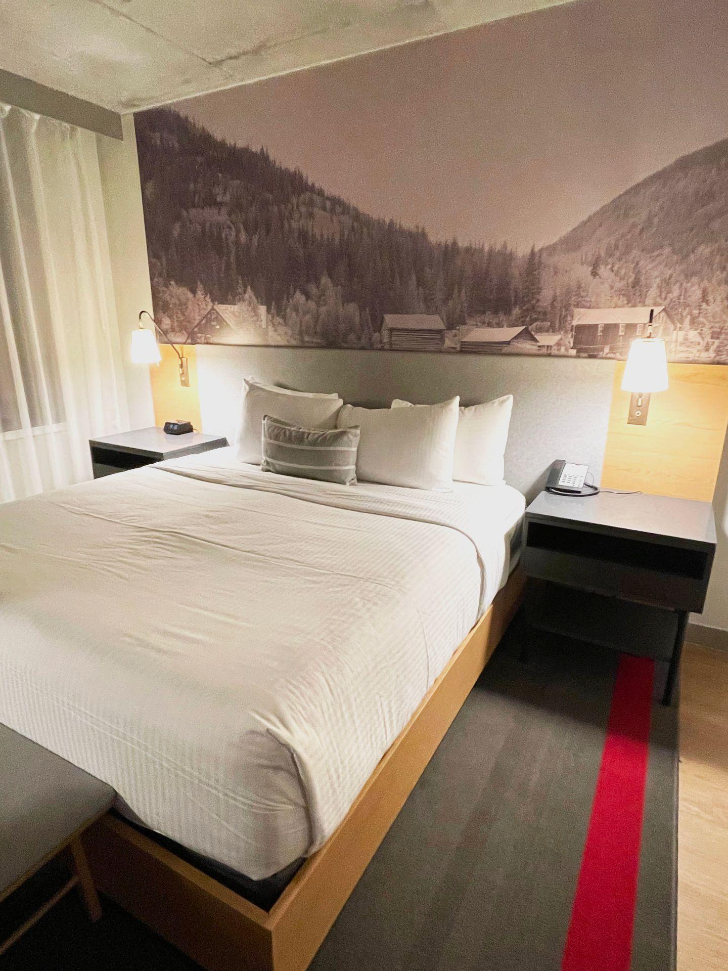 A hotel room with a neatly made bed and a grayscale mountain photo on the wall behind the bed.