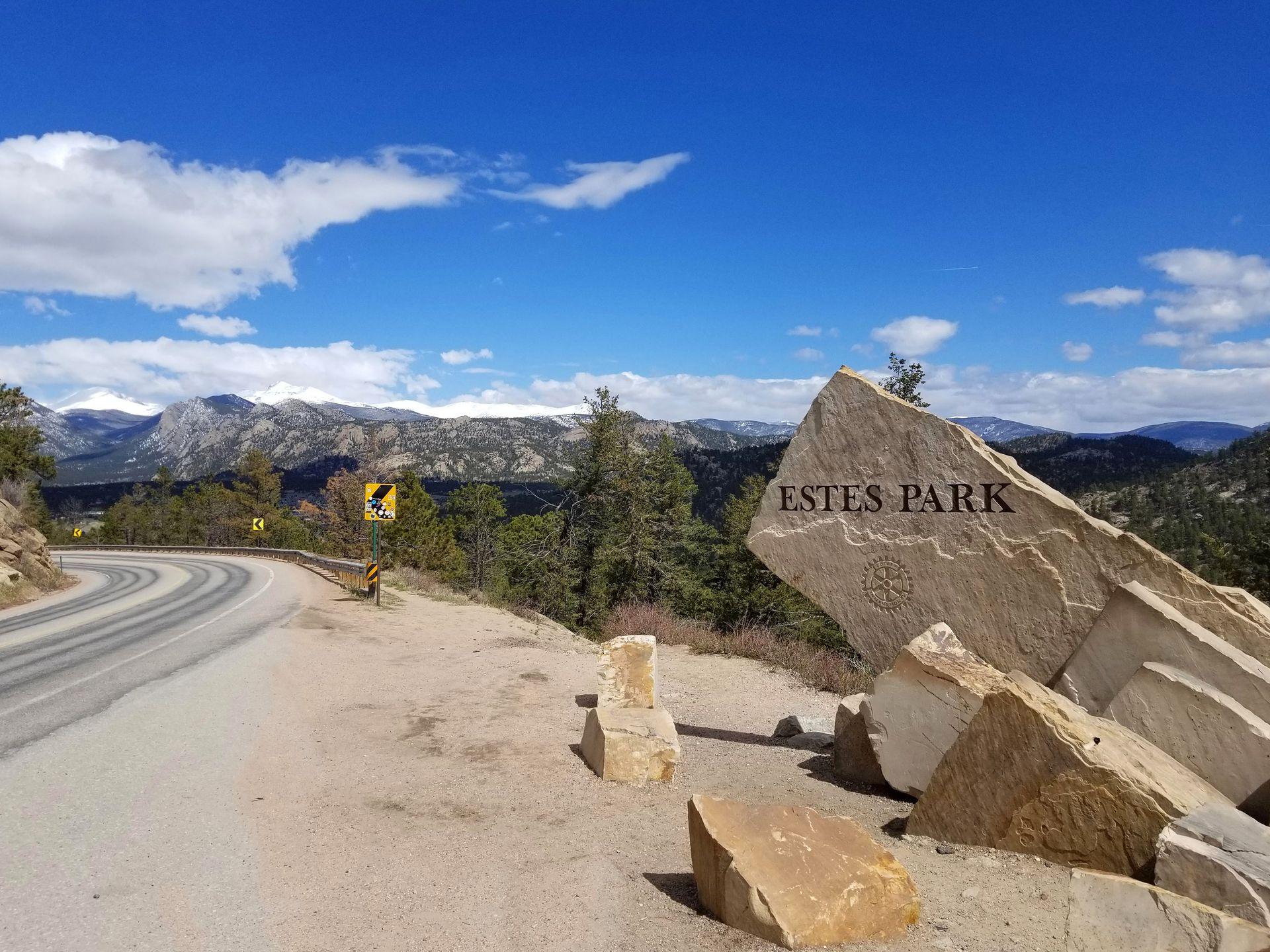 A sign made of huge stones that says "Estes Park." It is next to a windy road and there are mountains in the distance.