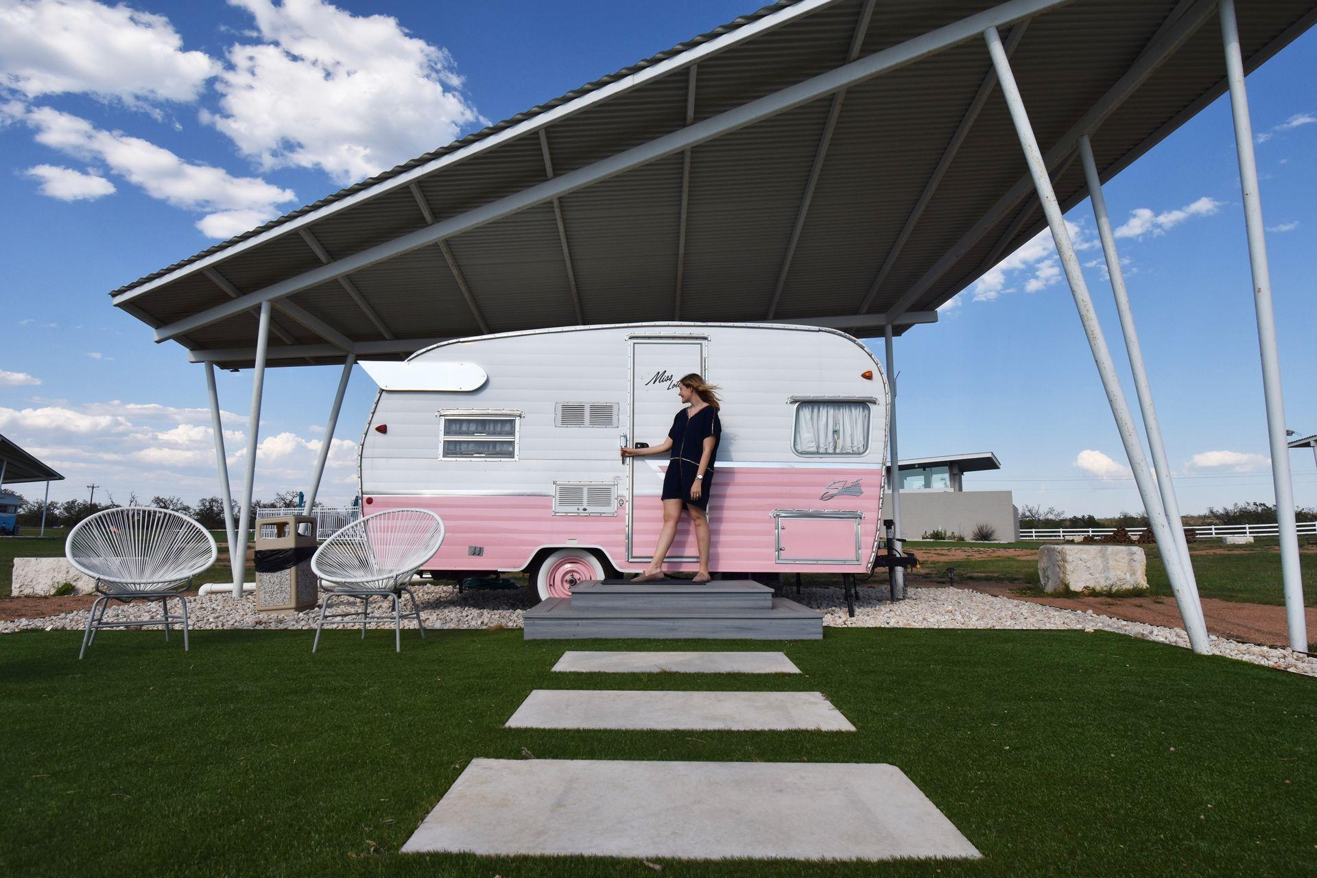 Lydia standing in front of a pink and white trailer at Blue Skies Retro Resort. She wears a blue dress and is holding the door.