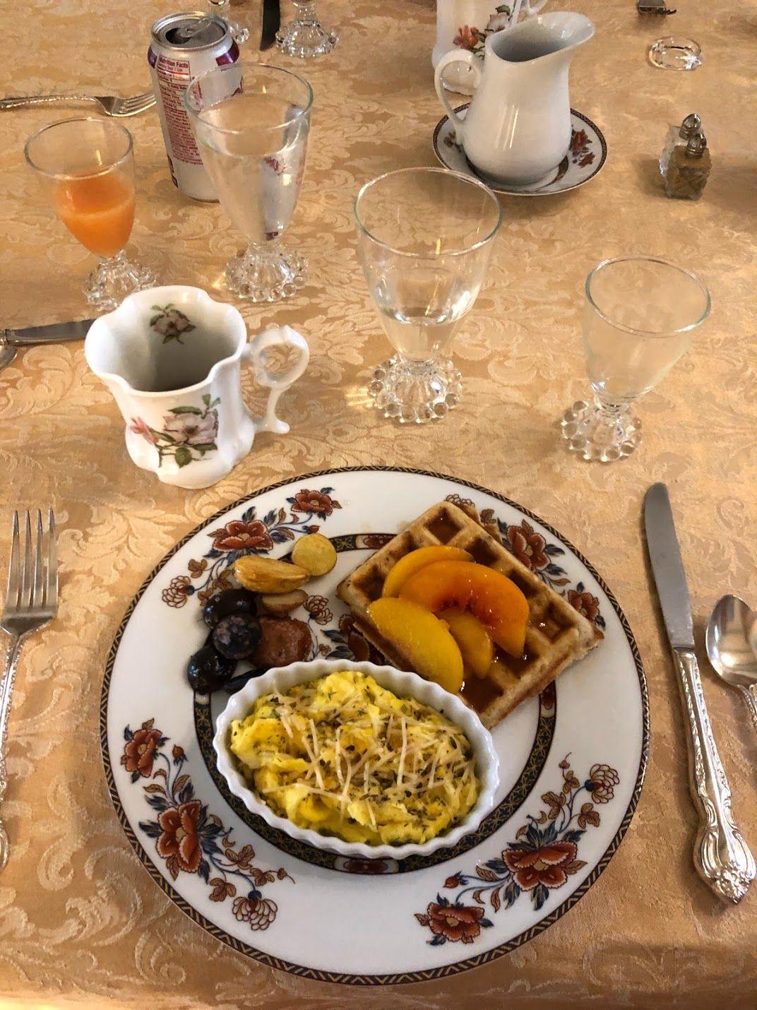 A plate of breakfast food with a waffle topped with fruit and cheesy potatoes. There is beautiful china and a mug next to the plate.