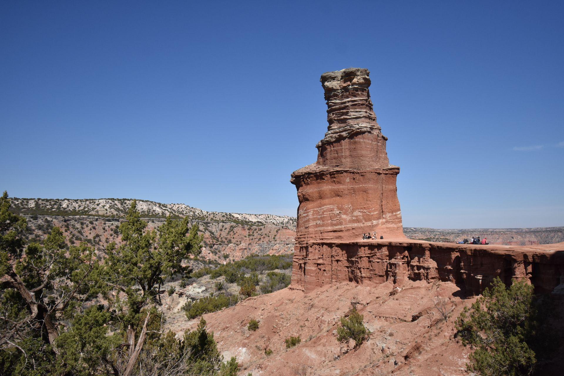 A large orange rock formation that resembles a lighthouse. There are some below it and a view of the canyon in the background.