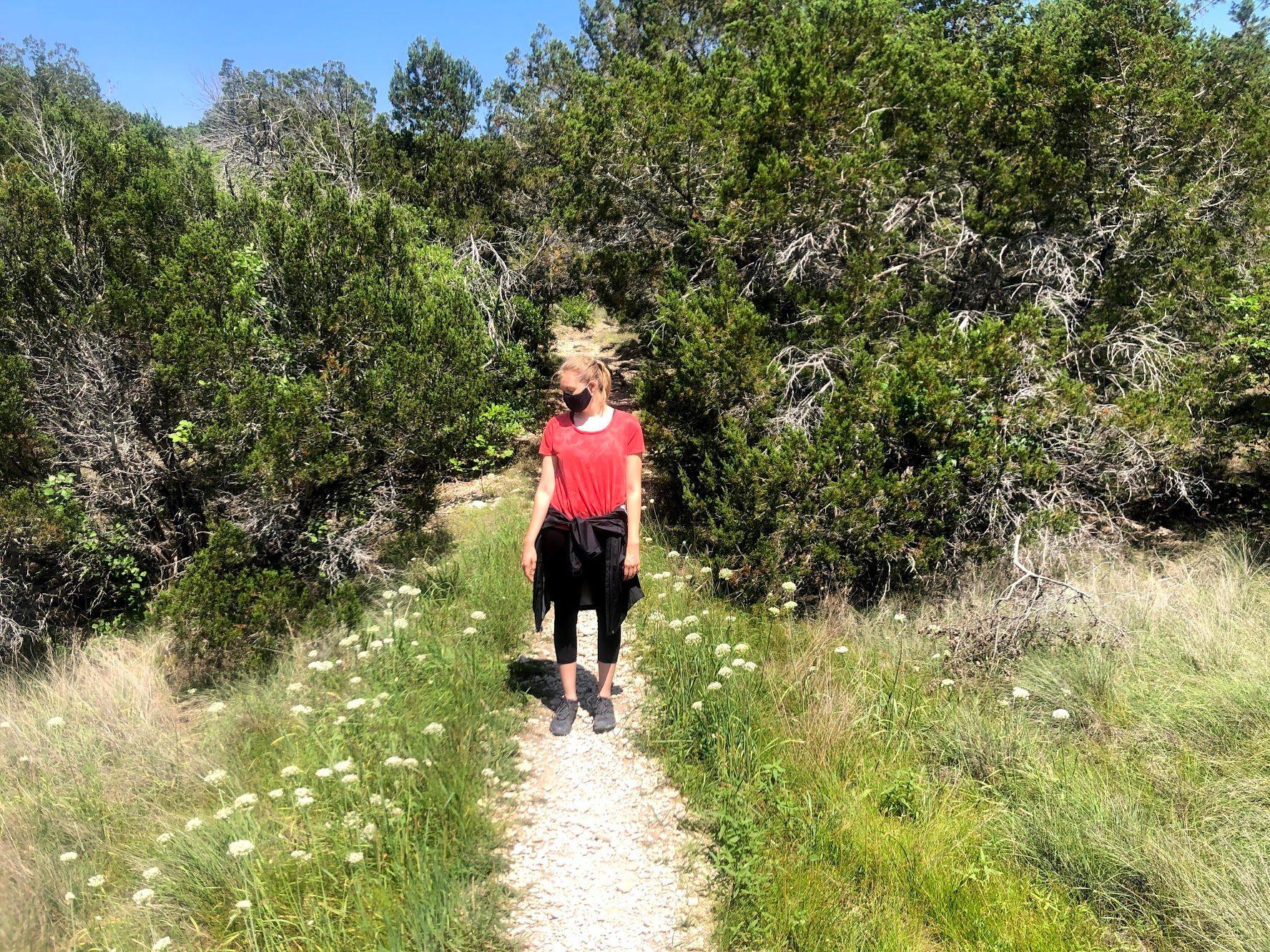 Lydia standing on a trail, with wildflowers on either side and trees behind her, wearing a mask.
