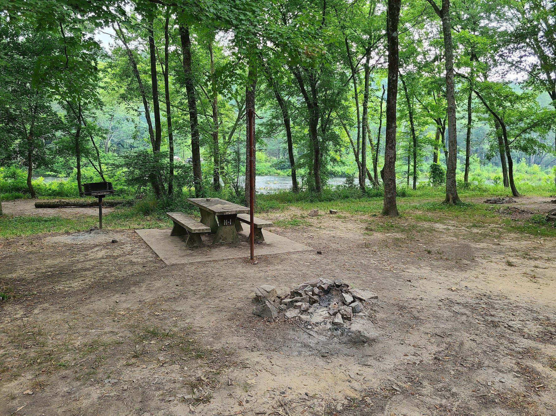 A campsite with a firepit, a picnic table and a grill. There are trees and a small peek at a river in the background.