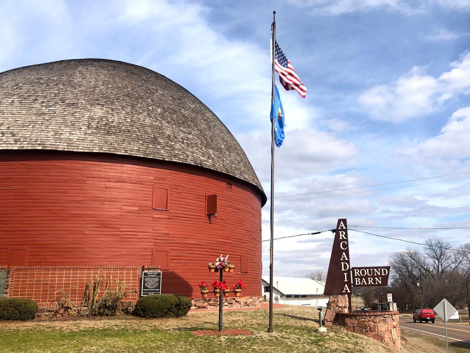 An orange, round barn with a rounded brown roof. There is a sign for Arcadia Round Barn and an American flag.
