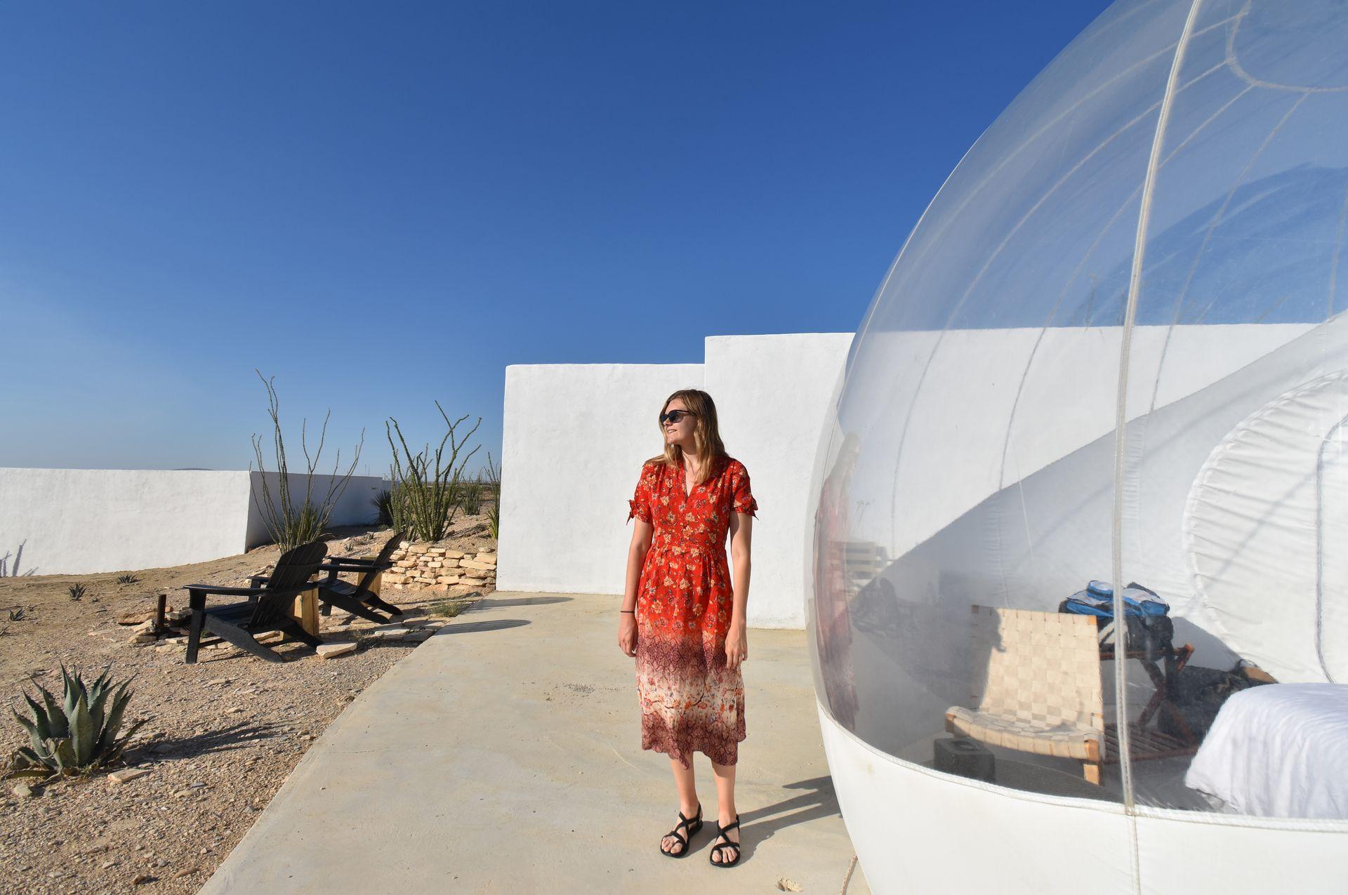Lydia wearing a red dress standing next to a clear bubble. There are white walls and some outdoor chairs in the background.