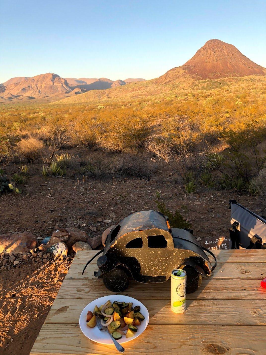 A picnic table with a mini volkewagen beetle, a plate of food, a can of white claw and mountains in the background.