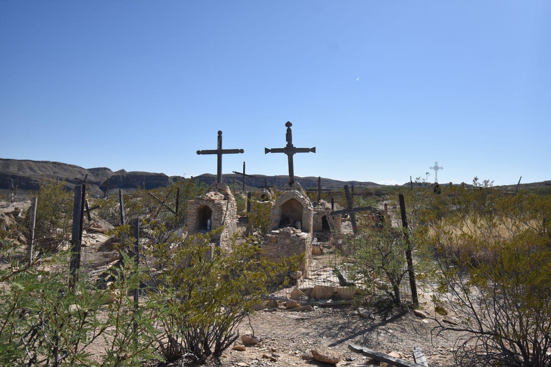 Crosses and a couple gravestones stick up out of the ground in the Terlingua Cemetery. They are surrounded by desert plants.