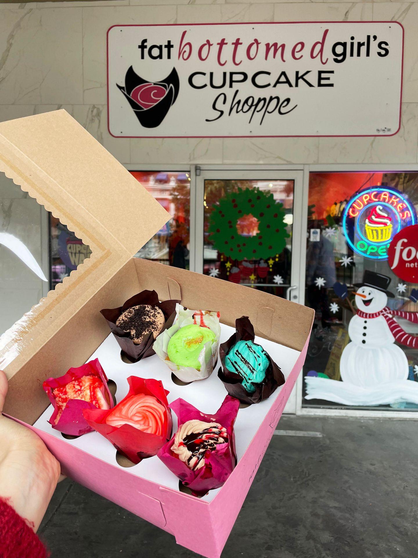 A box of six cupcakes being held up outside of Fat Bottomed Girls Cupcakes. The cupcakes have a mix of blue, green, brown, pink and red icing.