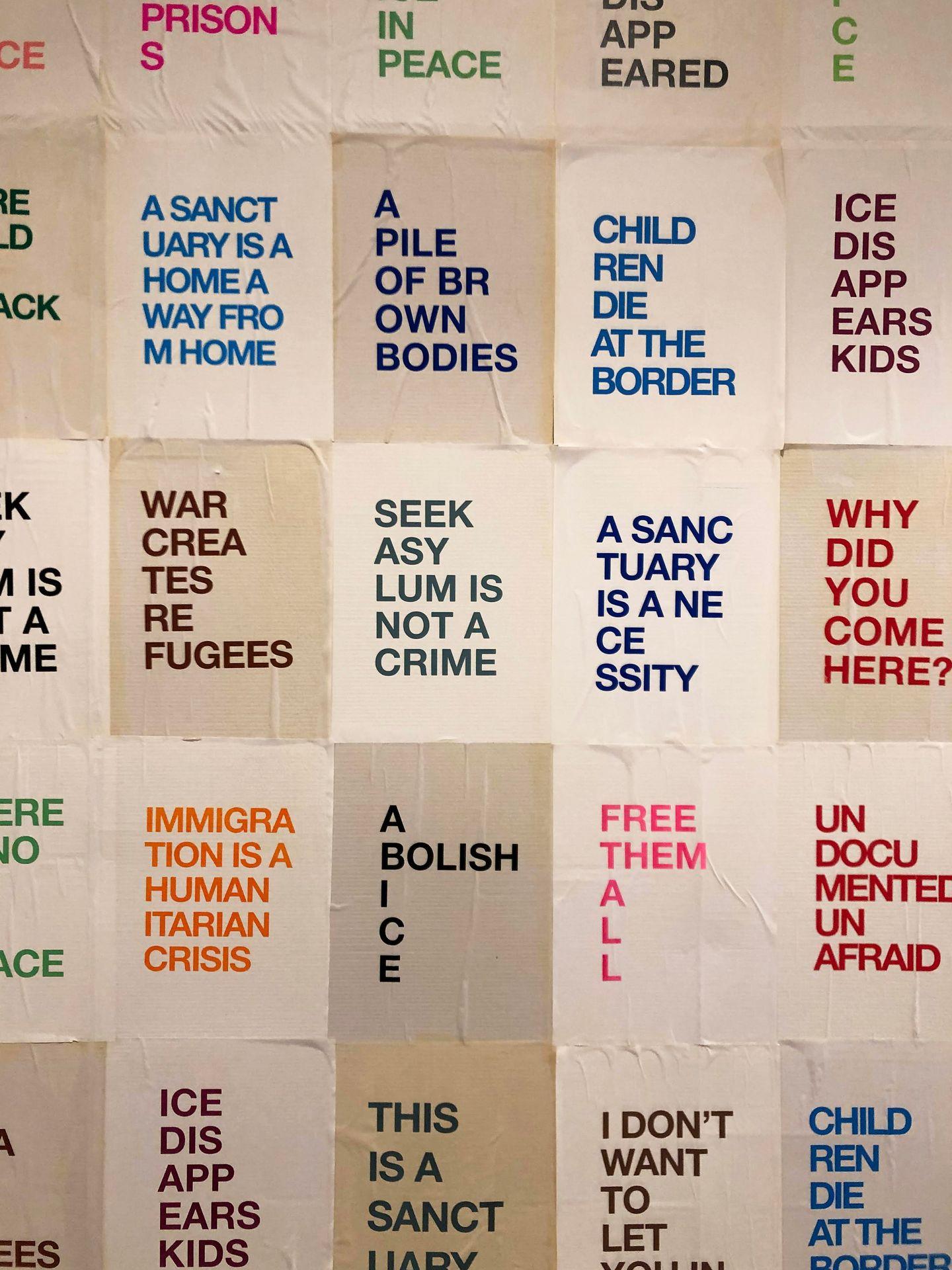 Several pages on a wall with words. Some read 'Free Them All,' 'Abolish ICE,' and 'A Sanctuary is a Necessity'