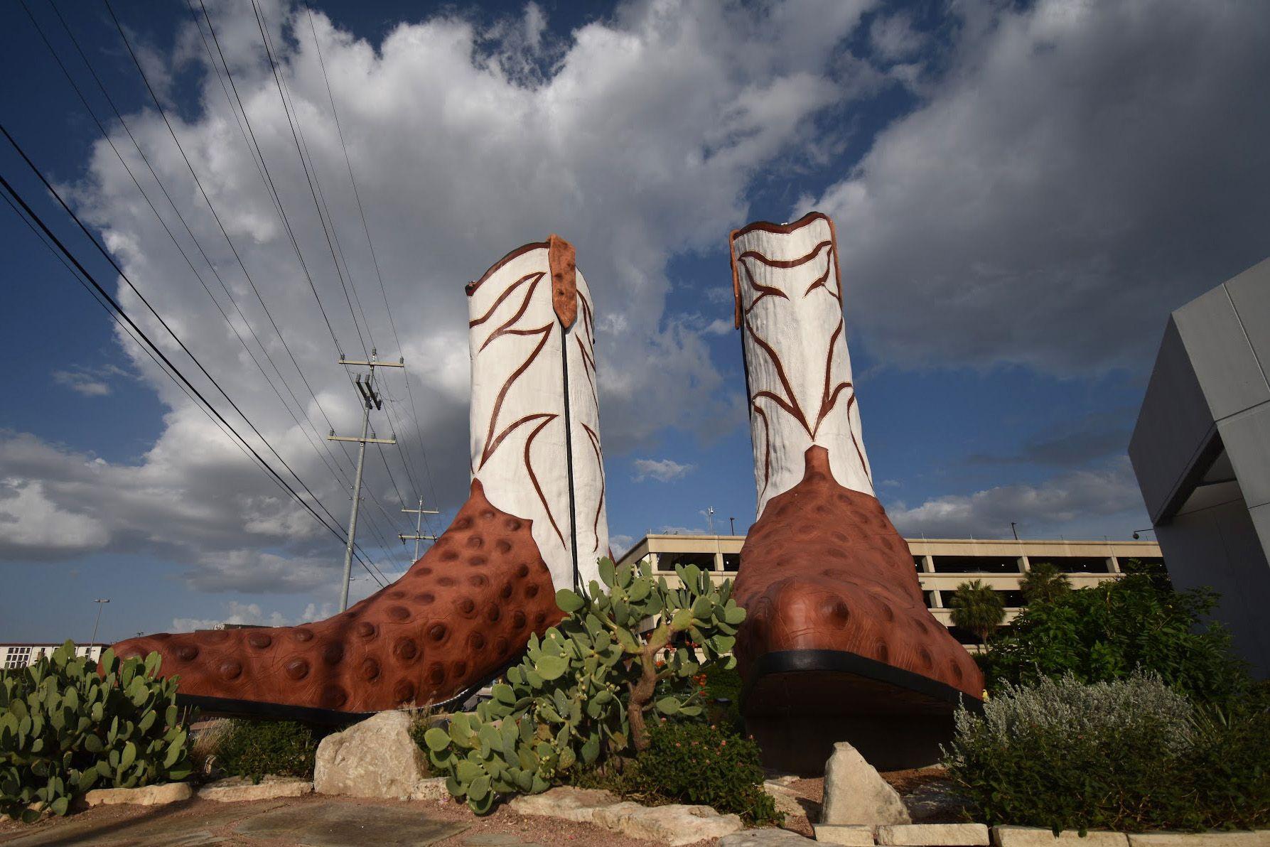 Looking up at two gigantic white and brown cowboy boots. There is a parking garage in the background.