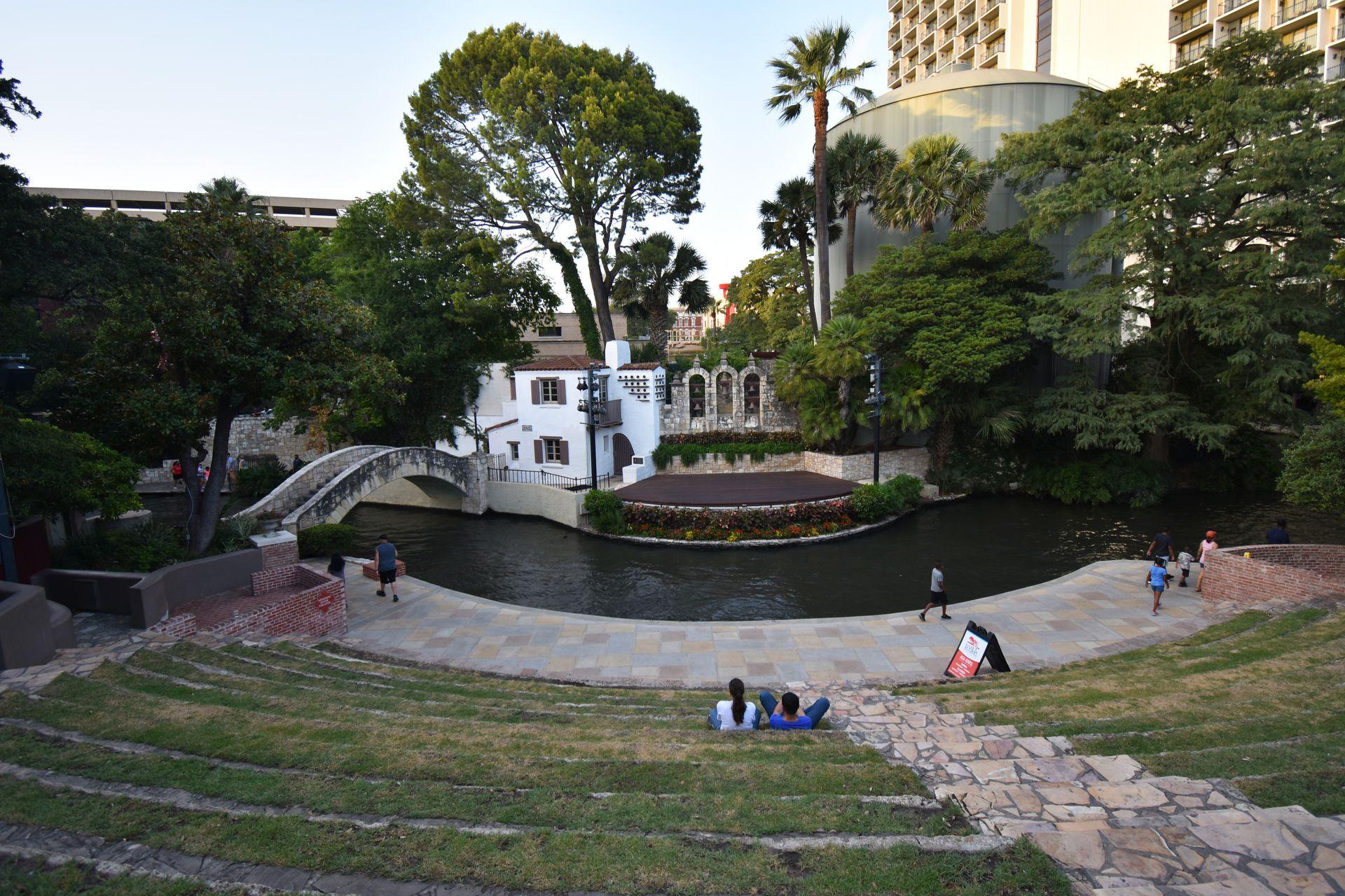 An amphitheater area along the San Antonio Riverwalk. There is a small bridge over the water and a white building across the river.