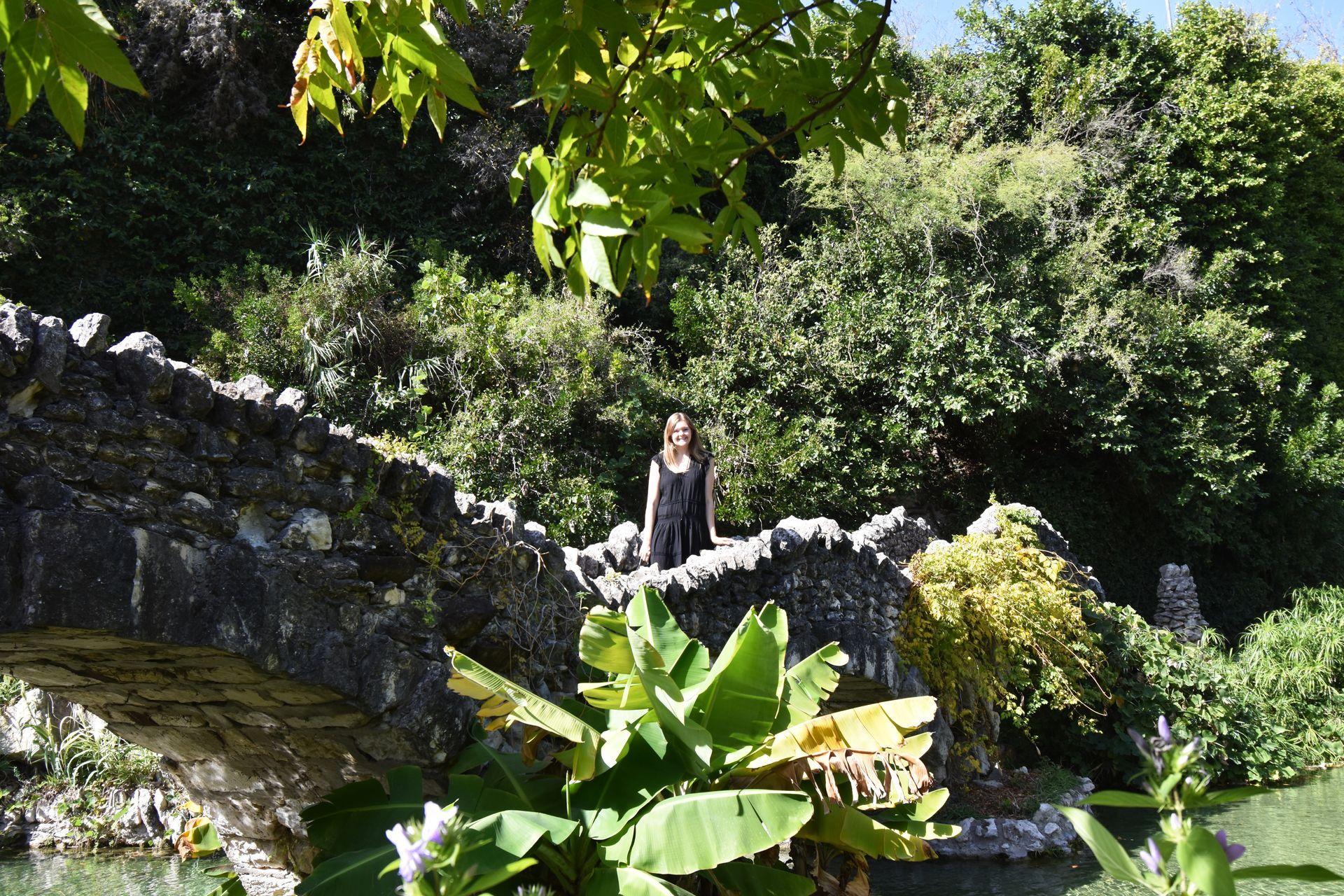 Lydia standing on a stone bridge in the Japanese Tea Garden. There are trees in the background and some large green leaves in the foreground.