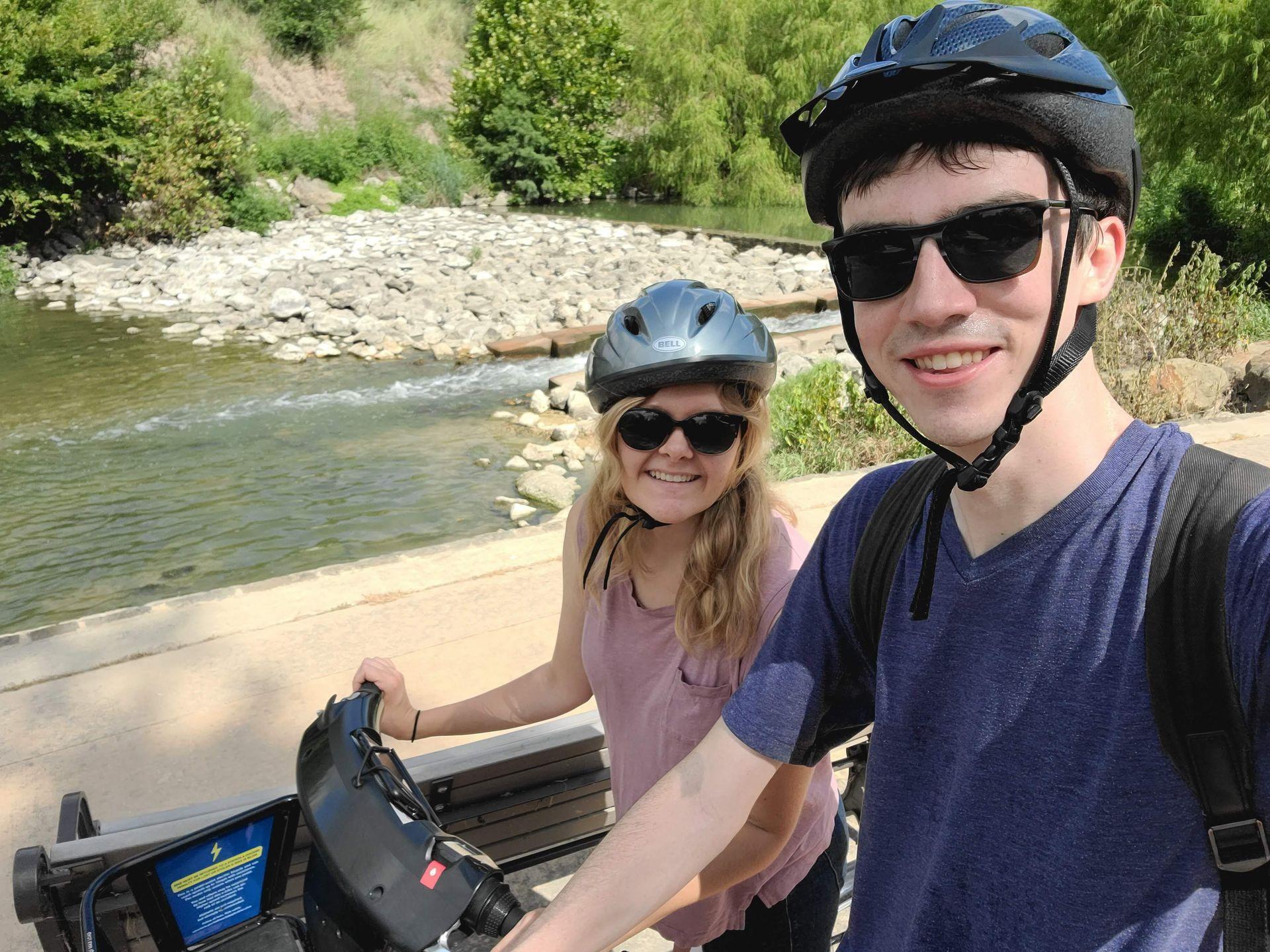 A selfie of Lydia and Joe sitting on bikes and wearing helmets. There is a river with rocks in the background.