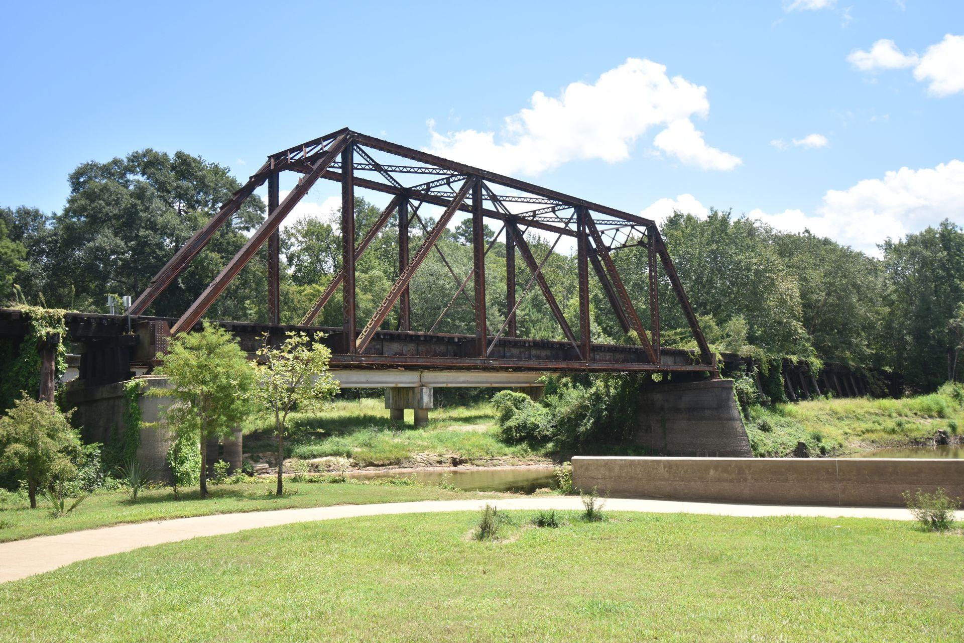 The Howe Truss Train Trestle Bridge from a short distnace away. It is surrounded by green grass and trees. At ground level below the bridge, there is a paved sidewalk.
