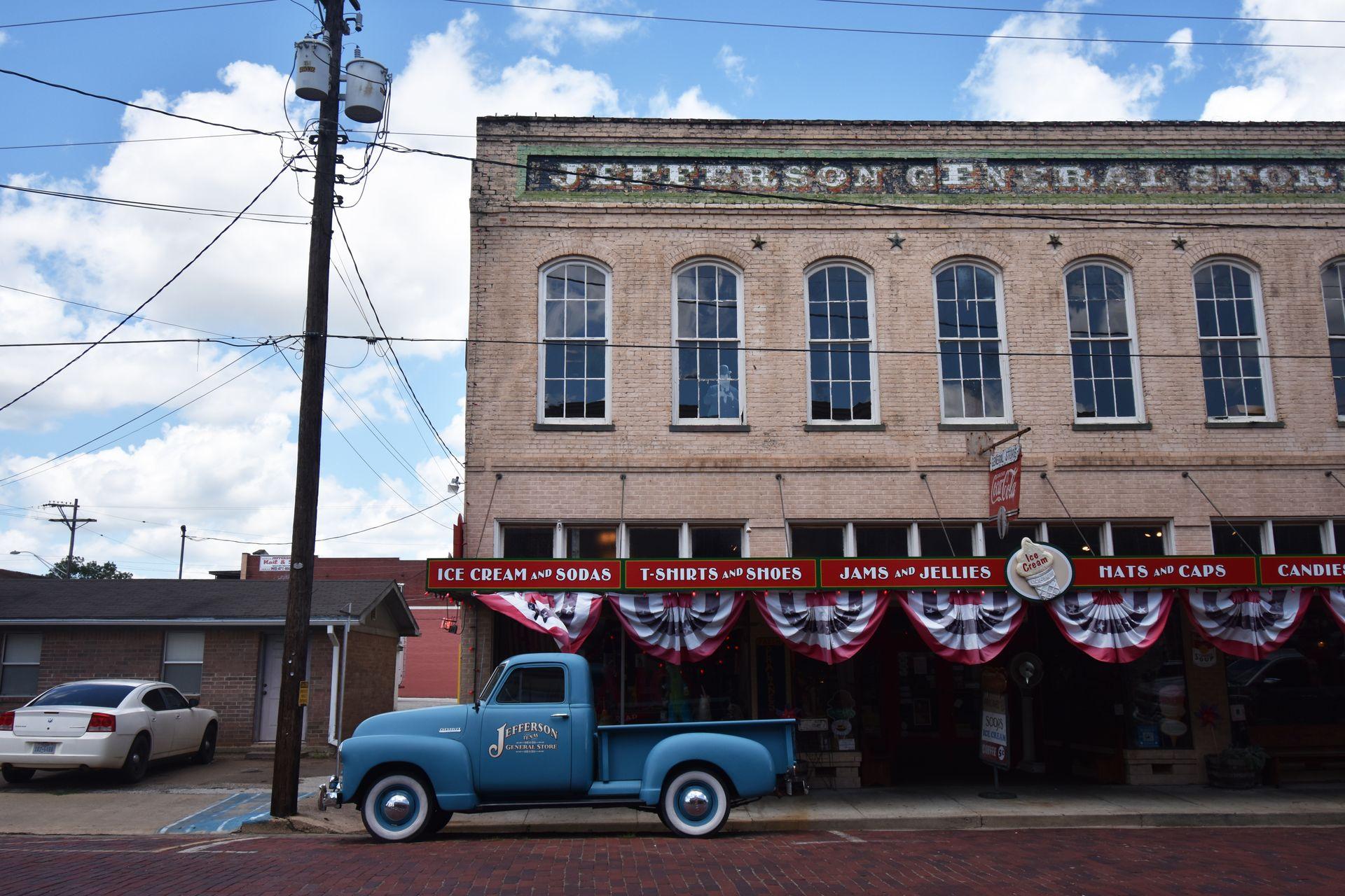 The exterior of the Jefferson General Store. There is a vintage blue pick up truck parked outside, red, white and blue awnings and signage saying "ice cream and soda," "t-shirts and shoes" and "jams and jellies"