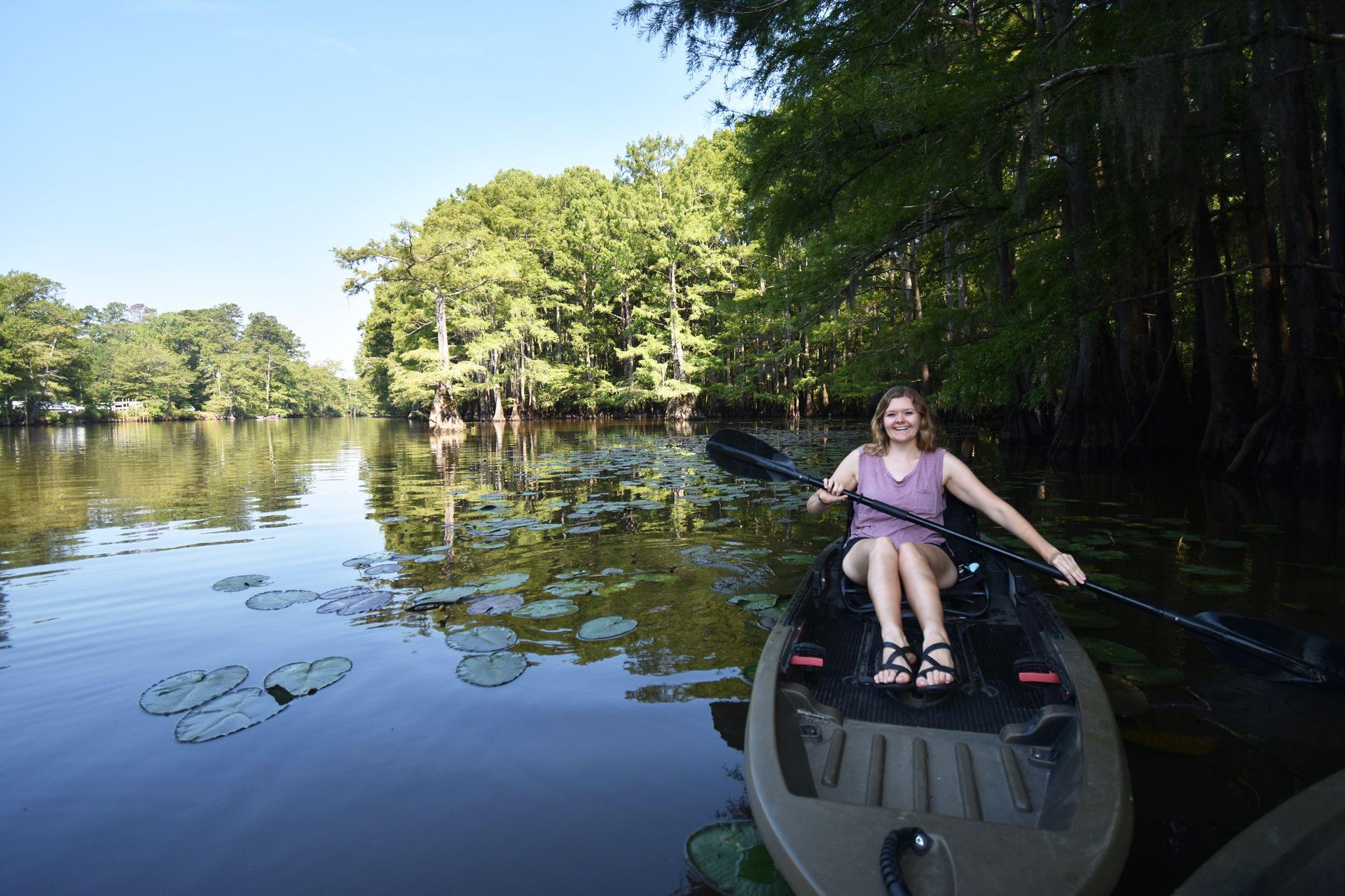Lydia sitting in a kayak on the Big Cypress Bayou. There is a large river area behind her with cypress trees along the shore.
