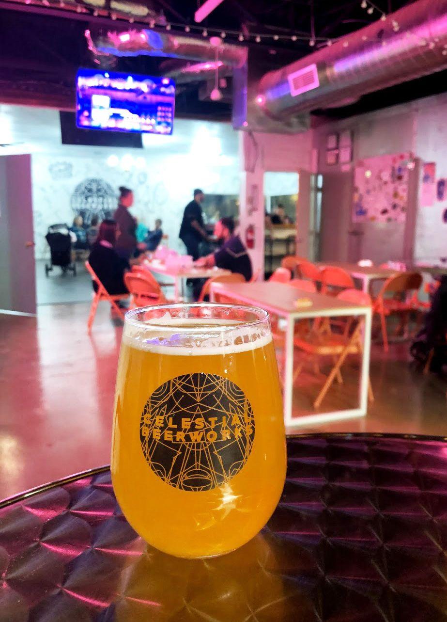A glass of light-colored beer at Celestial Brewery. The room is lit with pink lights.