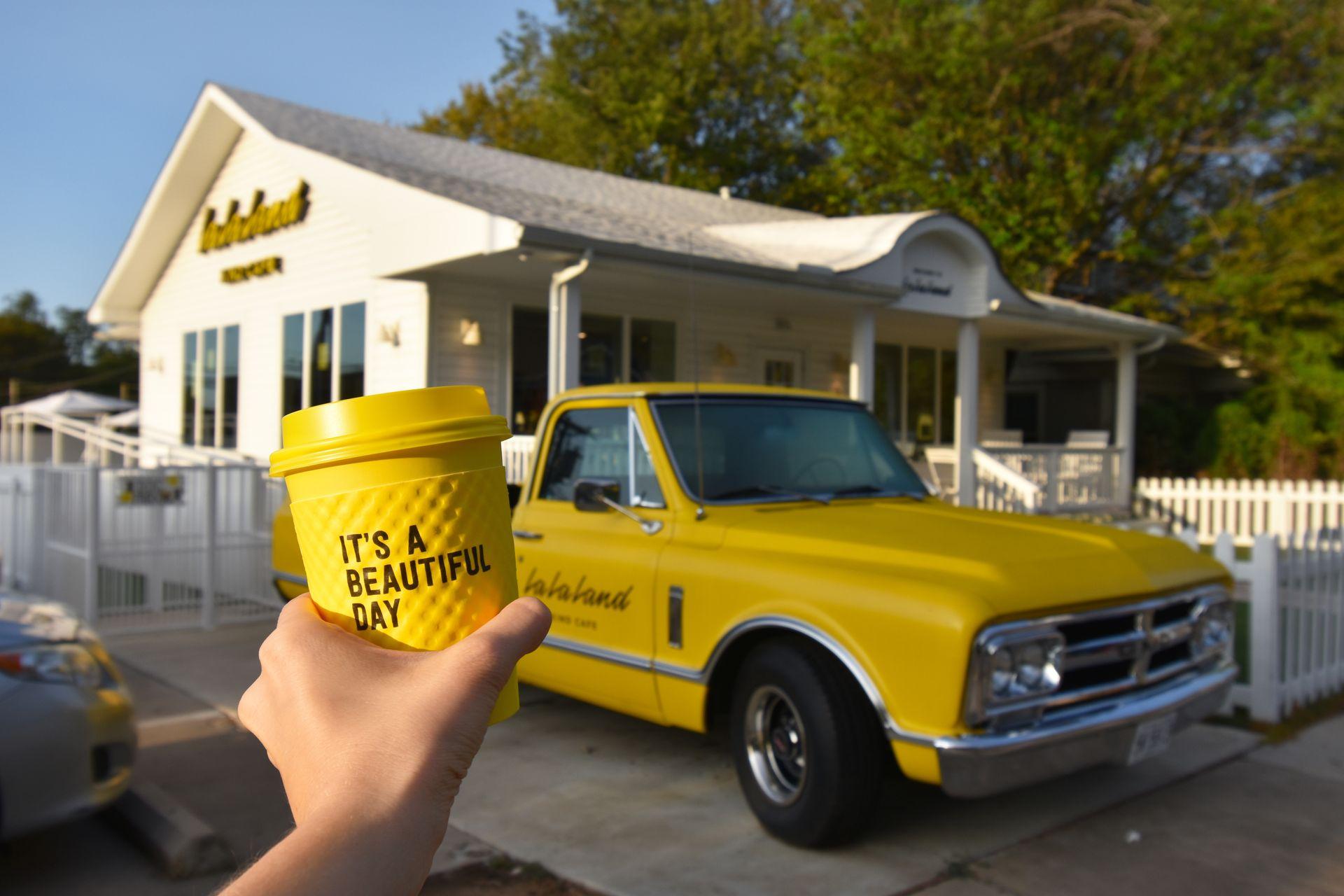 Holding up a yellow mug outside of La La Land Kind Cafe. There is a yellow pick up truck in front of the white coffee shop.