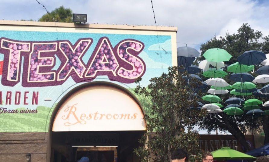A mural with the word Texas, with the letters filled with grapes, at the wine pavilion at the Texas State Fair. There are green, blue and white umbrellas hanging from the air to the right of the buliding.