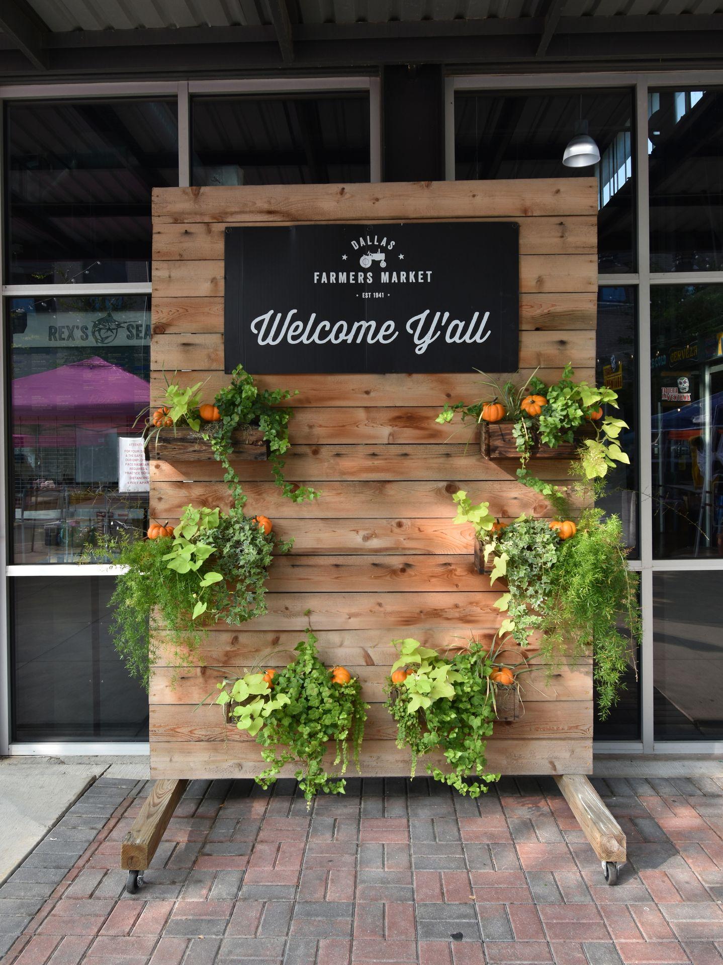 A wooden display that reads "Welcome Y'all" and has hanging plants outside of the Dallas Farmer's Market.