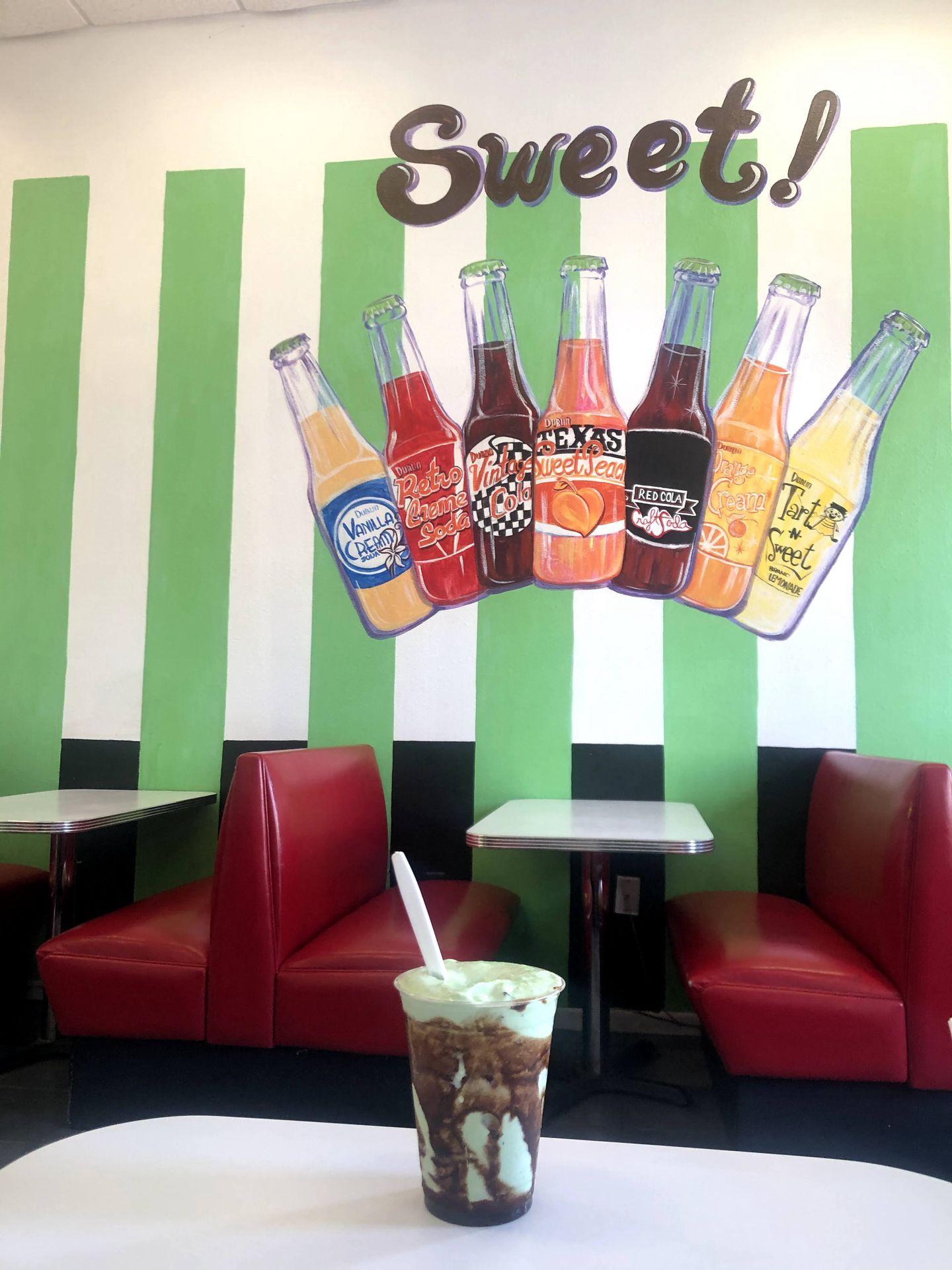 A green shade with chocolate syrup sits on a table in front of a mural that reads "Sweet" with several Dublin Soda bottles painted below it.