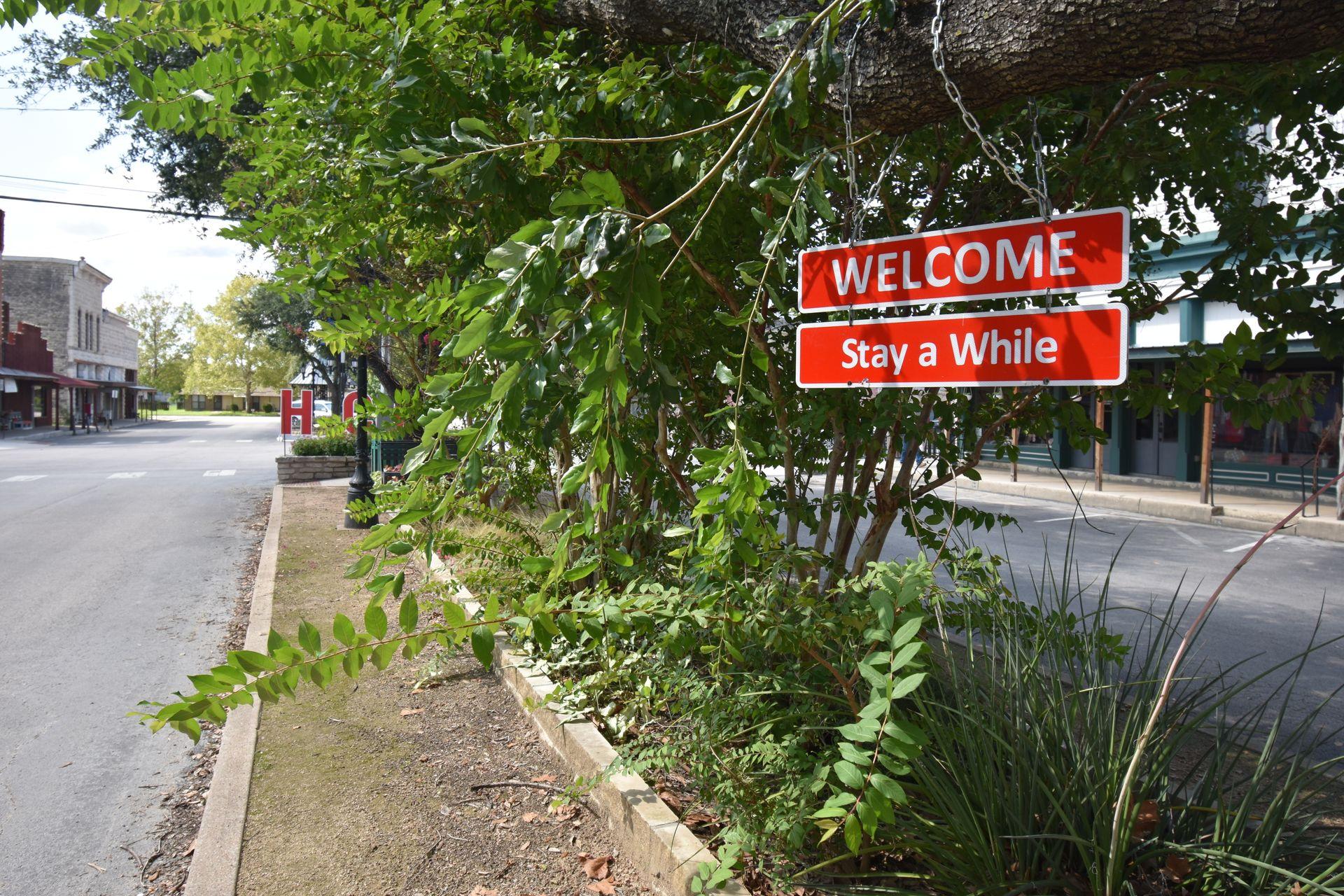 A red sign that reads "Welcome, Stay a While" among greenery in the middle of a road in downtown Hico.