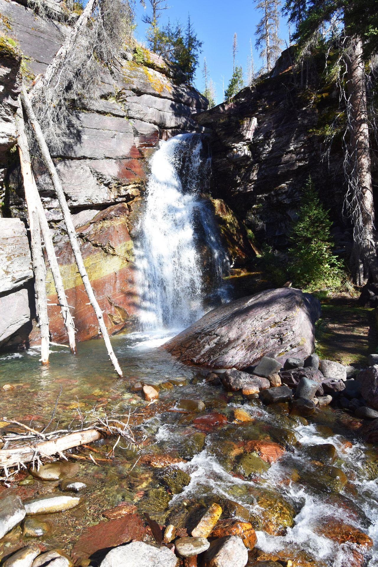 A waterfall coming down a rock wall. The wall is striped with red and yellow and there are red and yellow rocks in the water below.