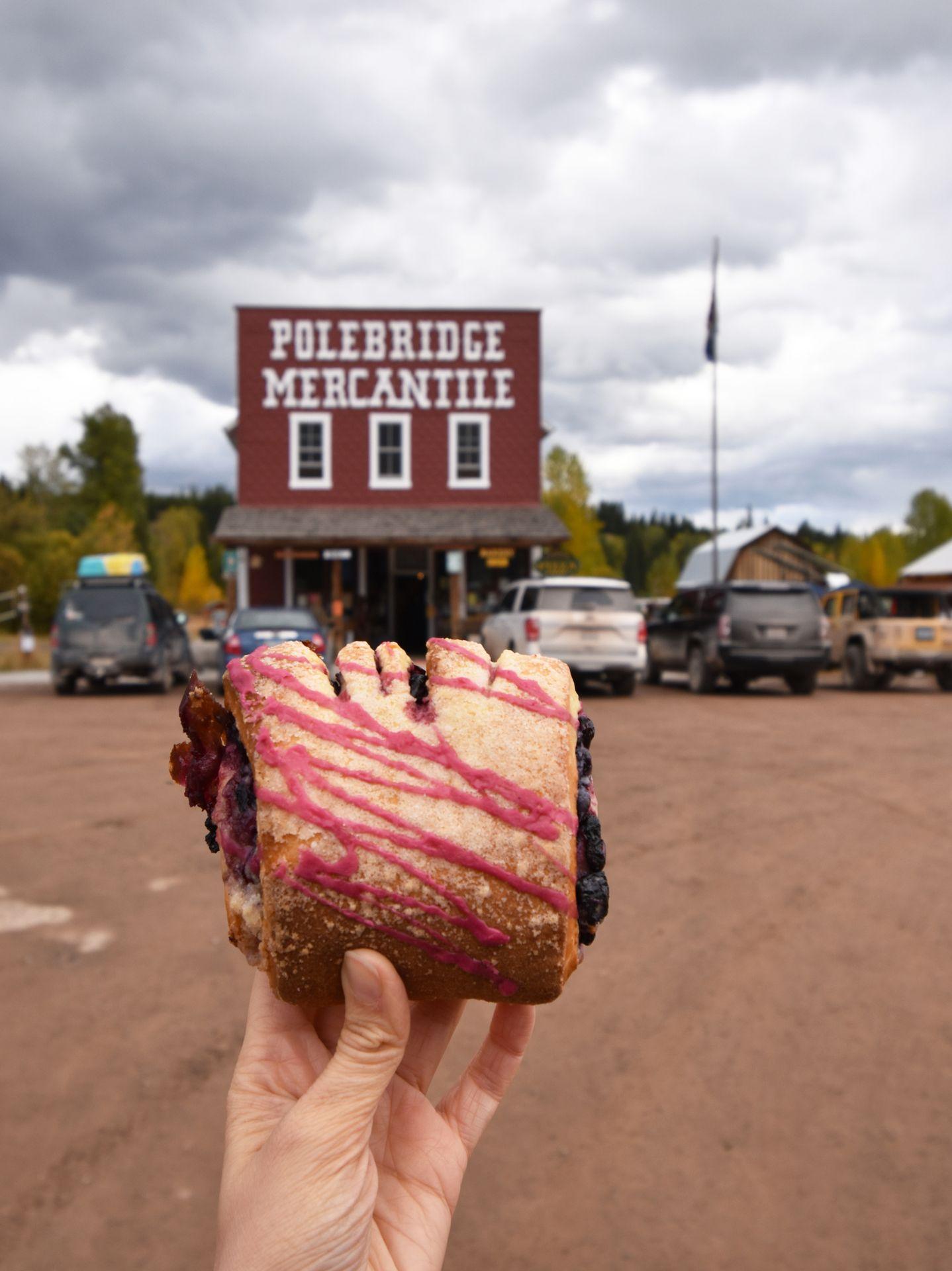 Holding up a huckleberry bearclaw in front of Polebridge Mercantile. The bearclaw has purple icing and Polebridge Mercantile is a red building with white letters.