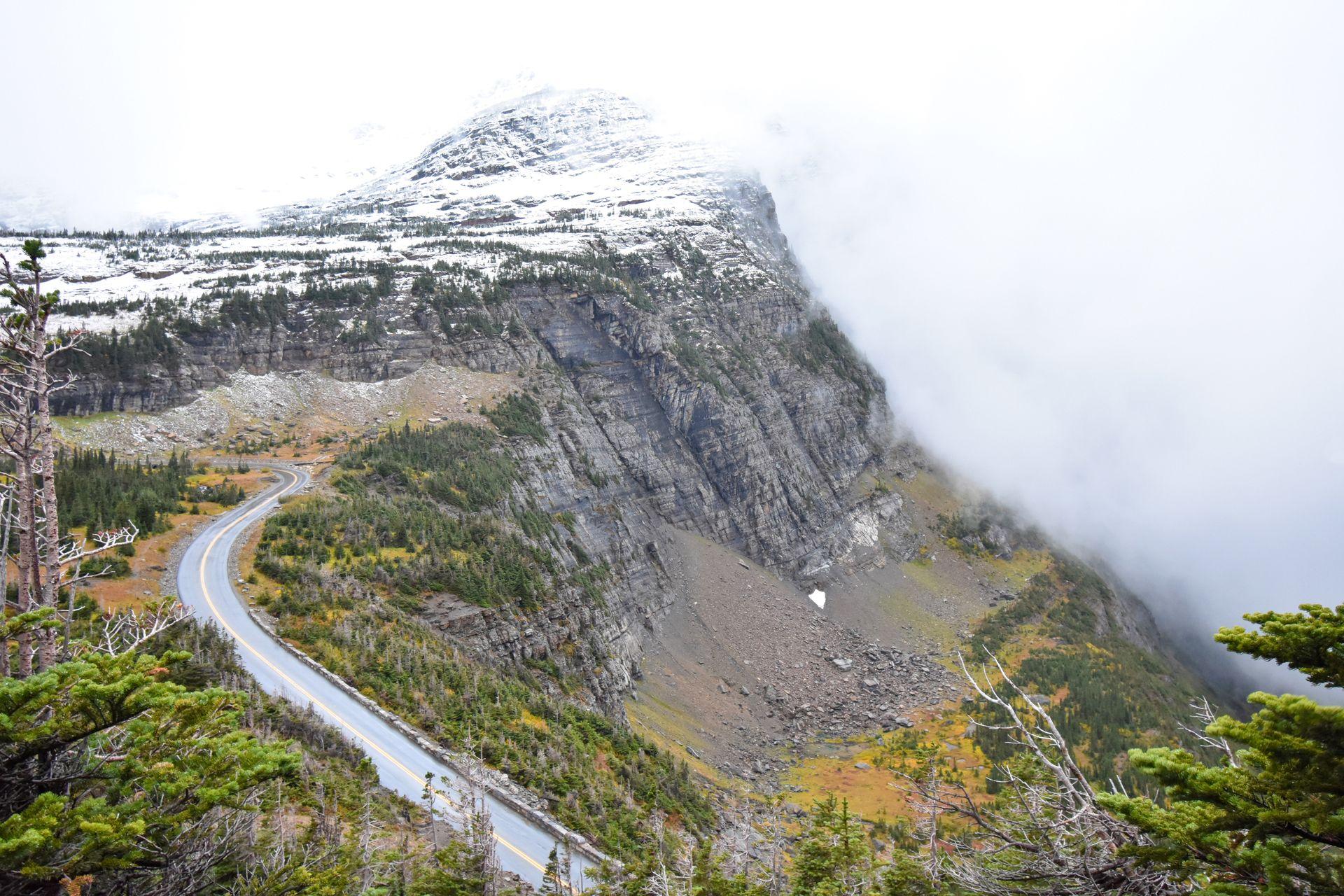 The Going to the Sun Road curving down a the side of the mountain. There is fog and snow on top of the mountain.